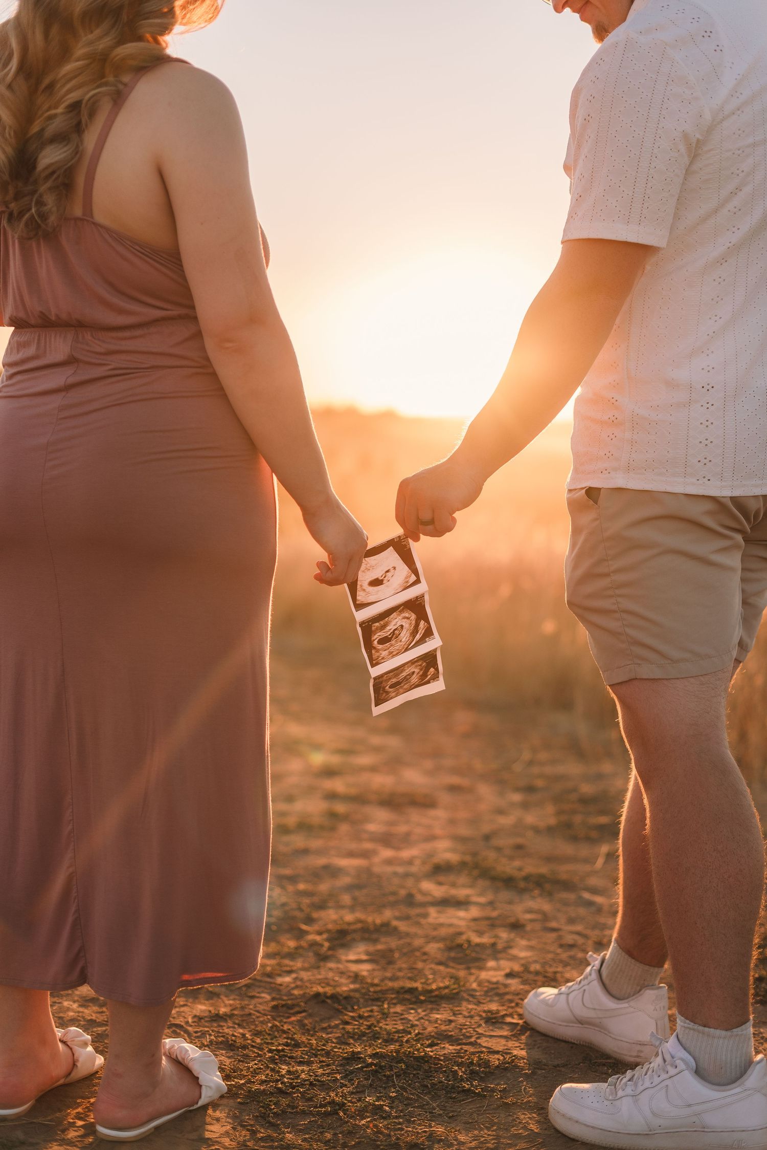 A pregnant person and their partner hold ultrasound photos at sunset in a golden-lit outdoor setting.