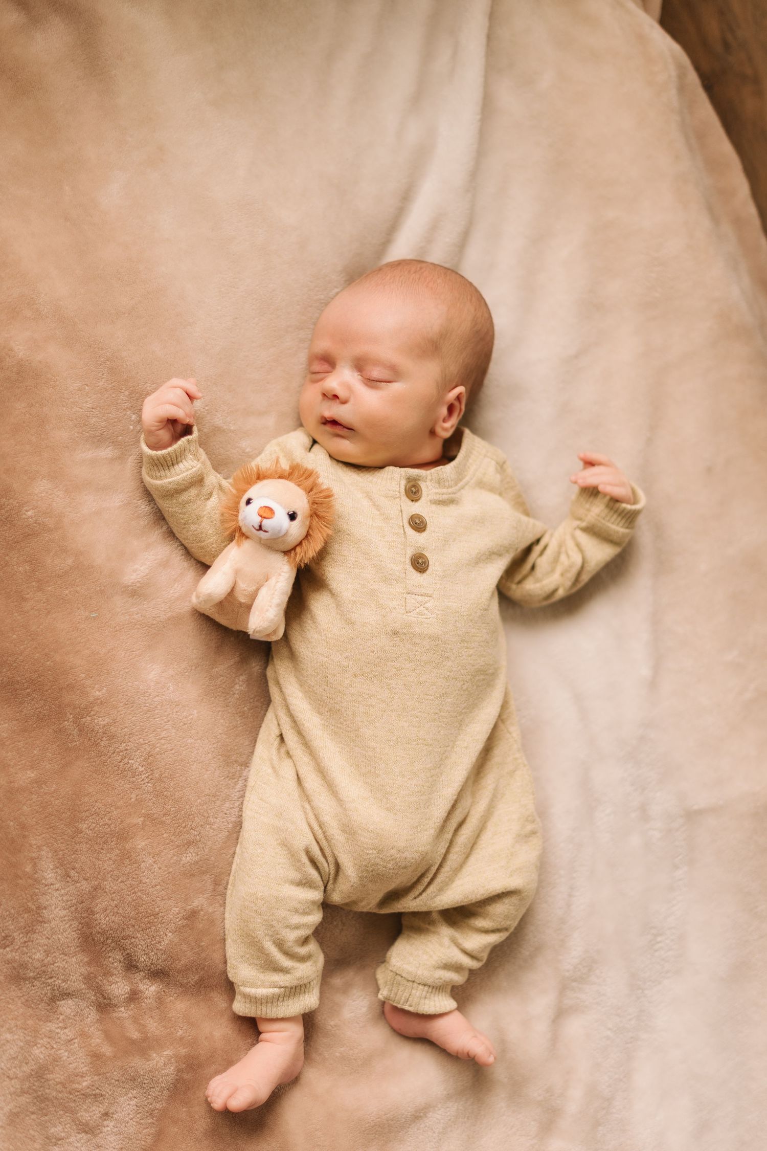 A baby wearing a beige onesie lies on a soft cream-colored blanket with a small plush toy companion.