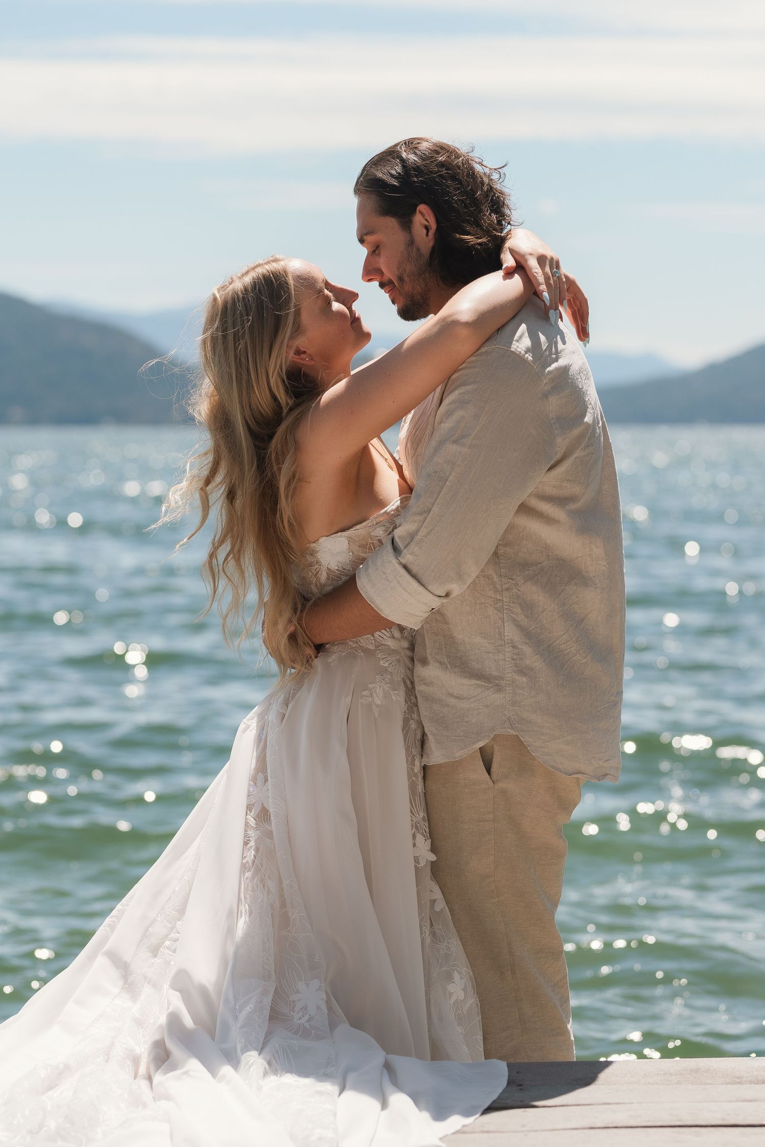 A bride and groom embrace on a wooden dock with sparkling blue water and mountains in the background.