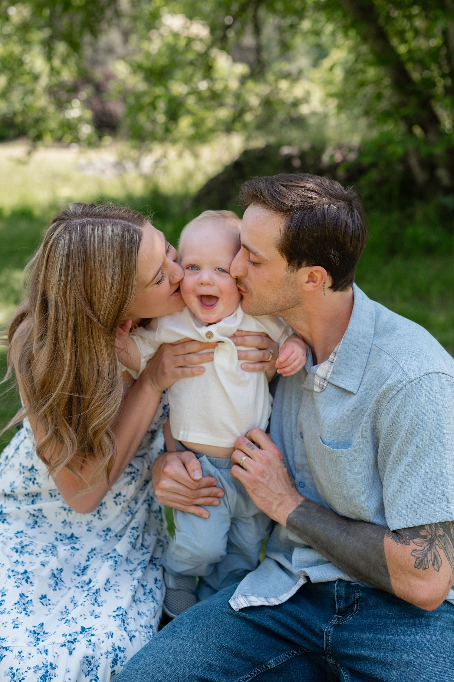 A family shares tender kisses and cuddles with a baby wearing white during an outdoor photo session in nature.