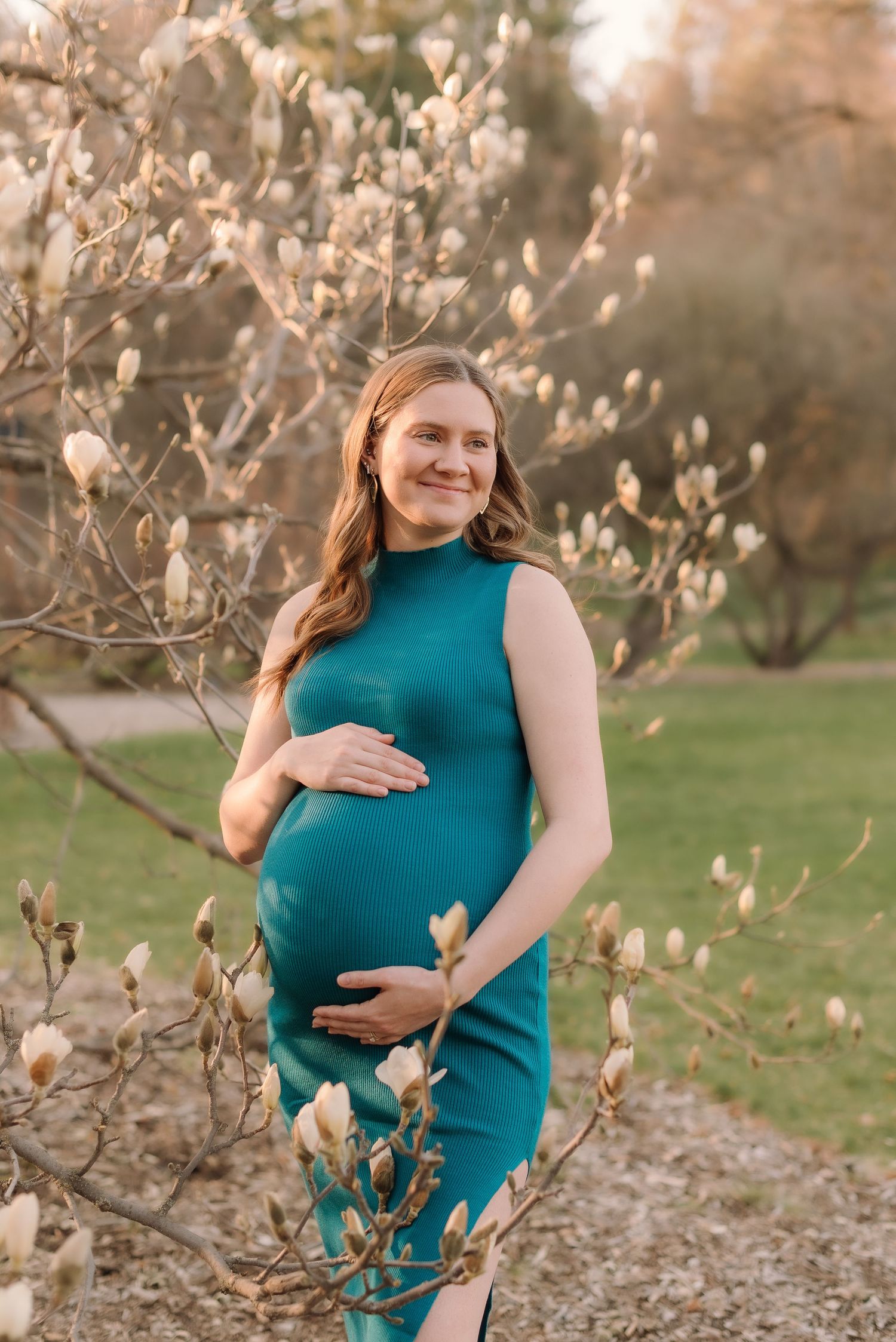 A woman in a teal dress poses for maternity photos among blooming magnolia trees during golden hour.