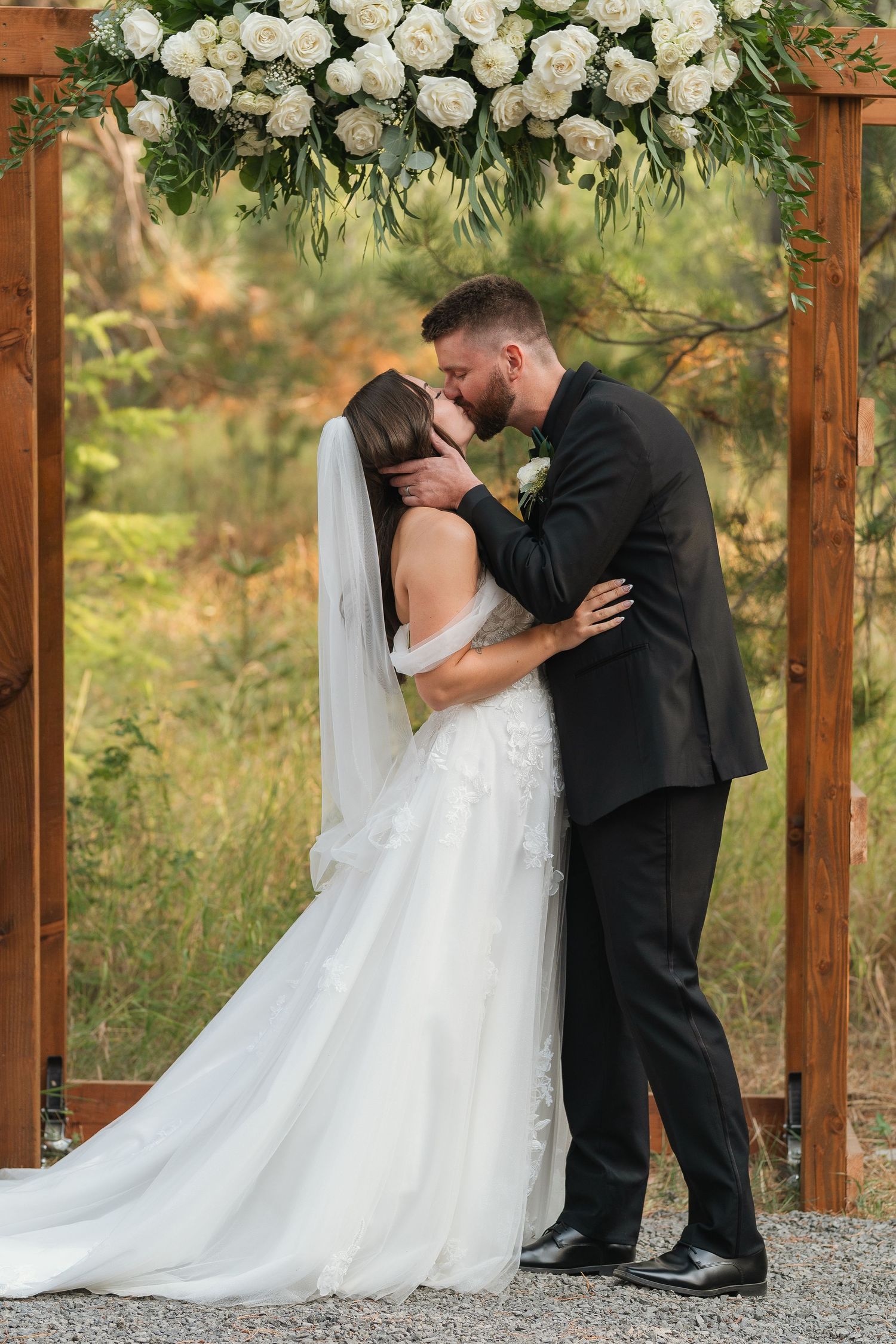 A romantic wedding moment under a wooden arch decorated with white roses and greenery in an outdoor garden setting.