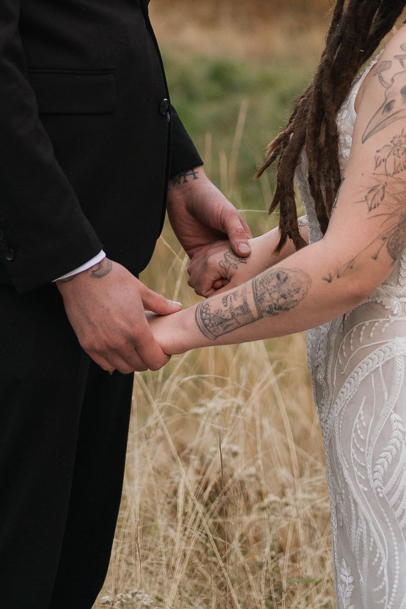 A close-up view of a couple holding hands outdoors, showing detailed lace from a wedding dress and tattoos.