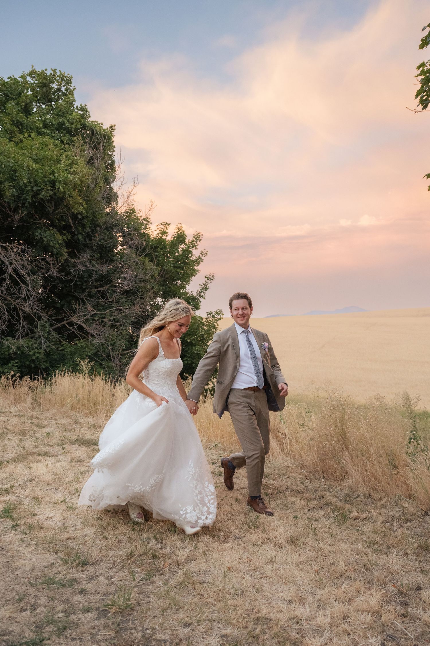 Bride and groom hold hands running through golden field at sunset in outdoor wedding portrait.