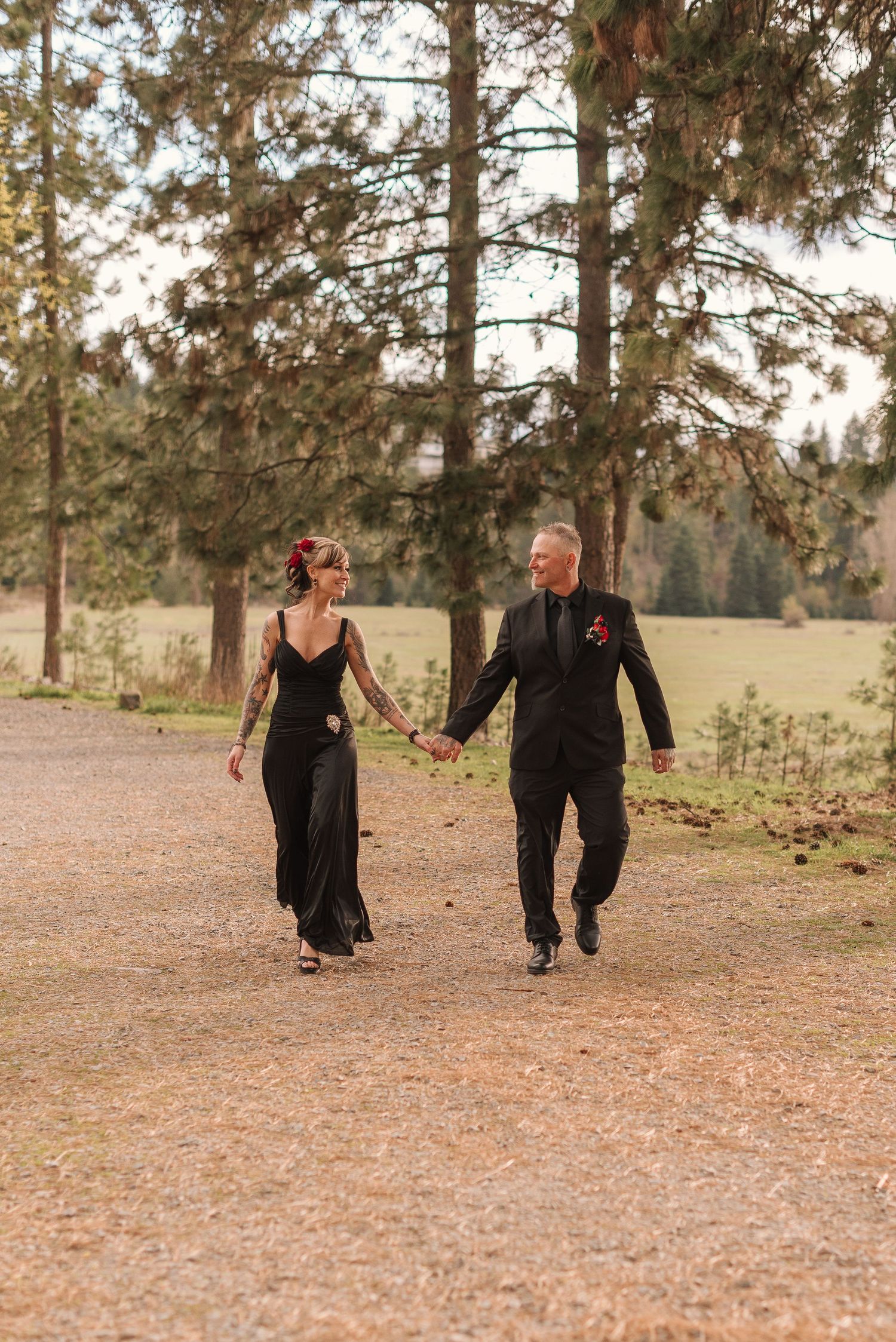A couple in black outfits walk hand in hand through a wooded area with pine trees in the background.