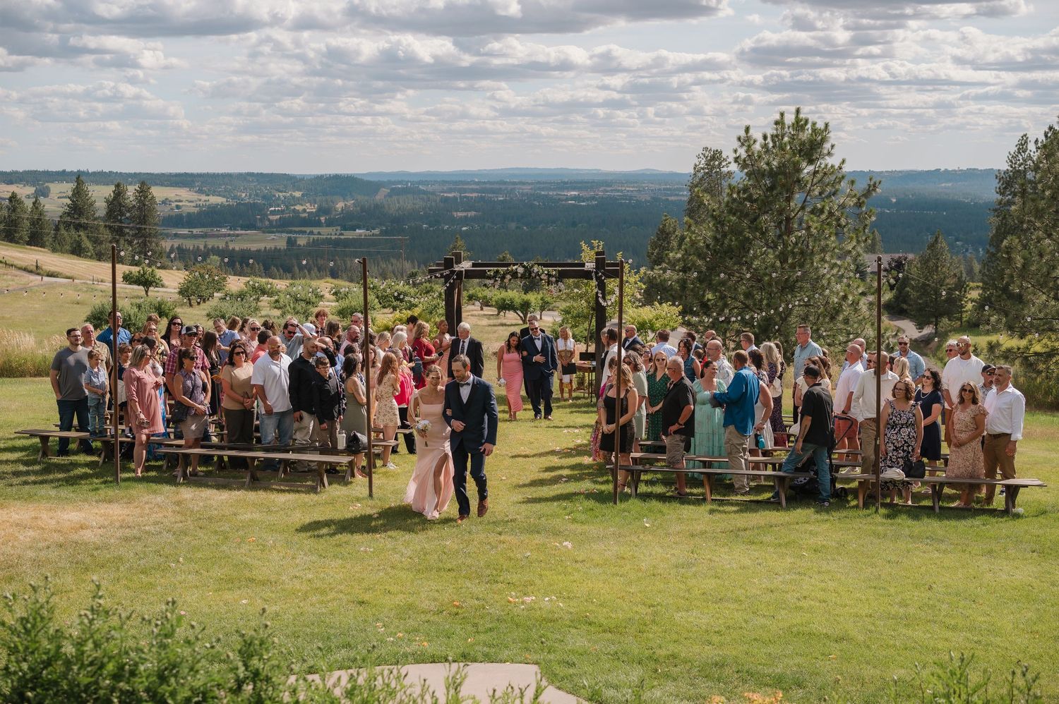 Outdoor wedding ceremony on green hillside with scenic valley views and wooden benches.