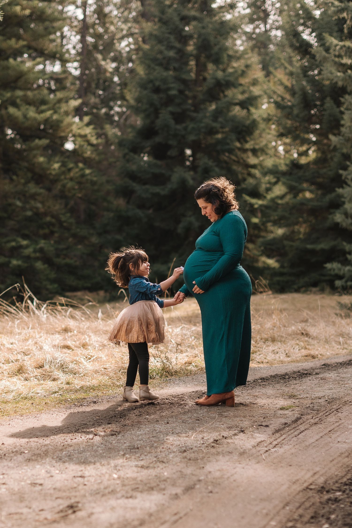 A parent and child share a tender moment on a dirt path surrounded by evergreen trees in golden afternoon light.