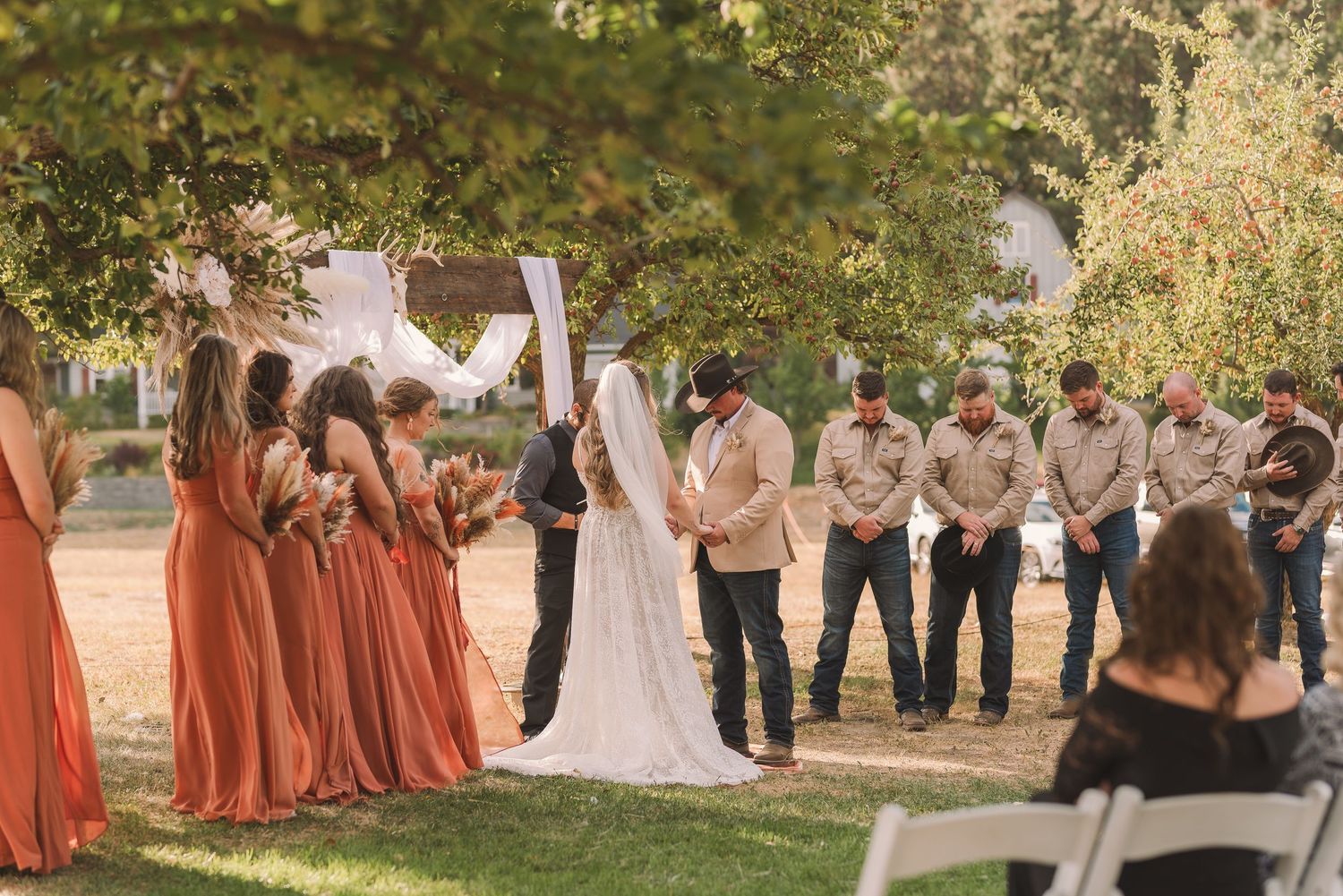 Wedding party and guests gather outdoors under trees with bridesmaids wearing coral-colored dresses.