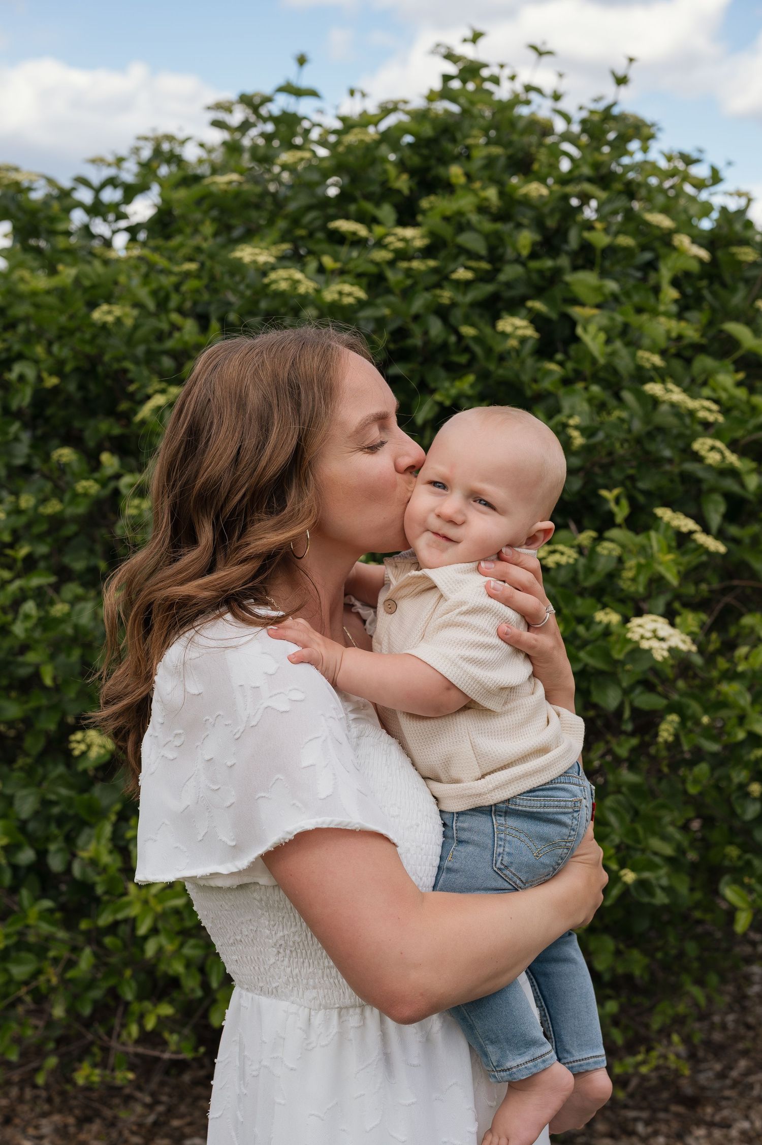 A mother in a white dress kisses her smiling baby boy outdoors with green shrubs in the background.