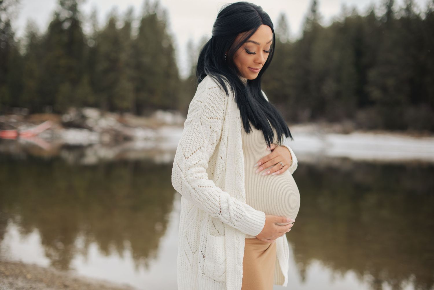Pregnant woman in white knit cardigan cradling baby bump outdoors near a serene lake with pine trees.