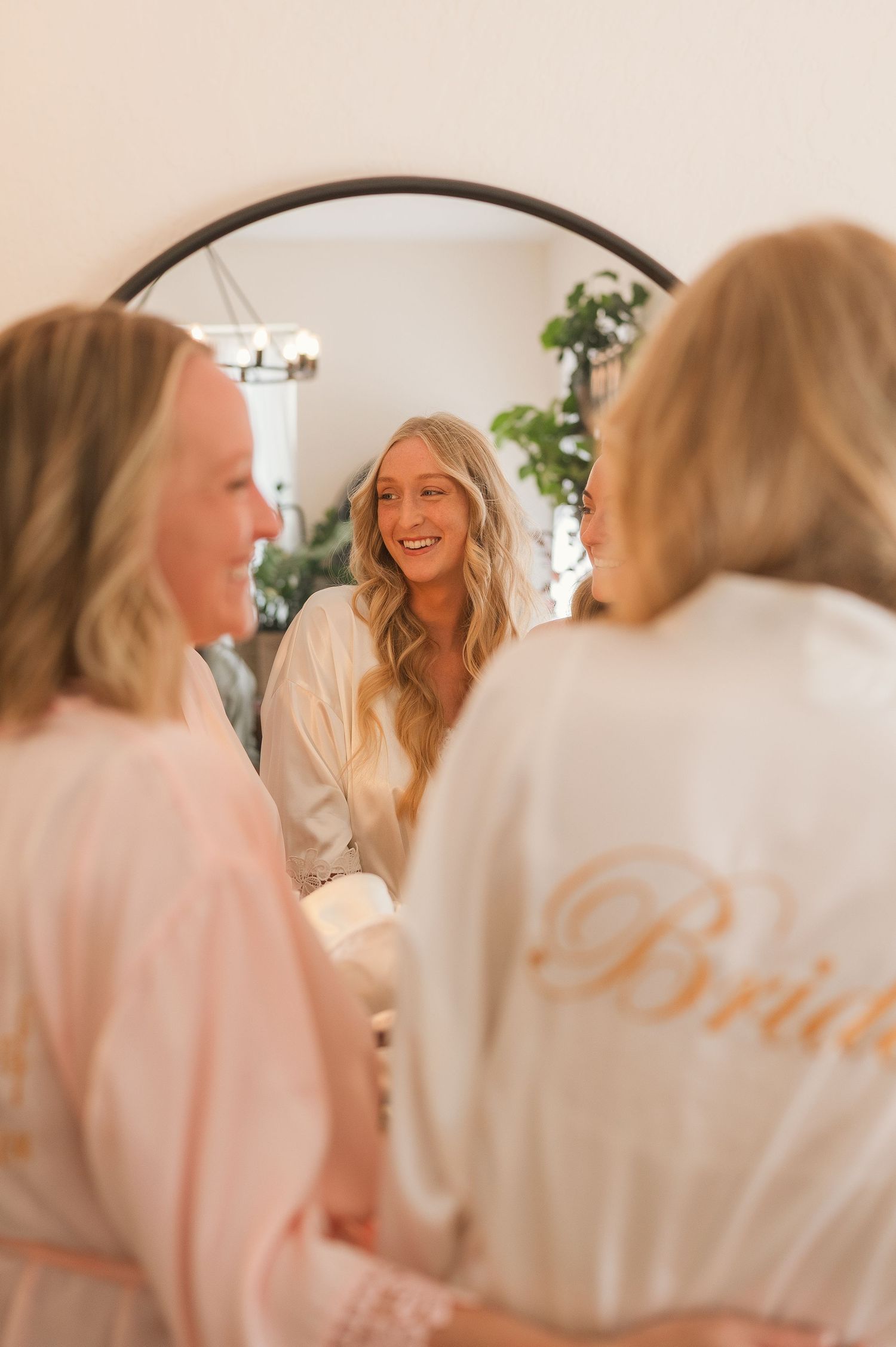 Bride in satin robe smiling at her reflection in a round mirror, surrounded by bridesmaids during wedding preparations.