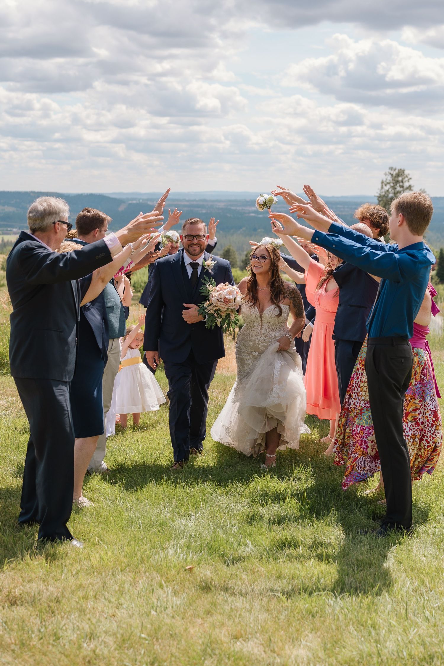 Wedding party and guests form a human tunnel in a grassy field under cloudy skies for a celebratory exit. Spokane Wedding Photography.