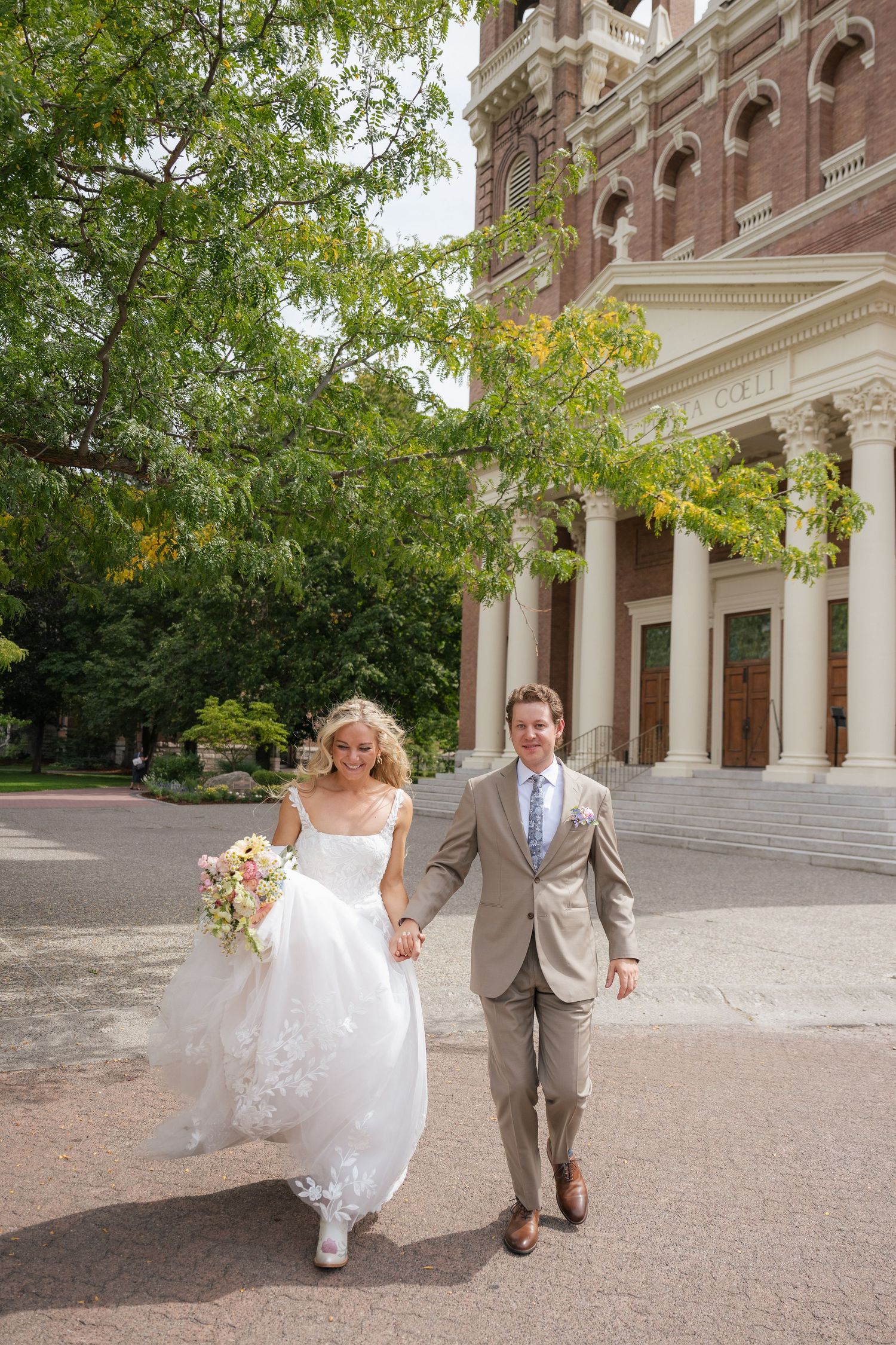 Bride and groom holding hands walking outside a historic church, bride in white lace gown holding bouquet, groom in tan suit.