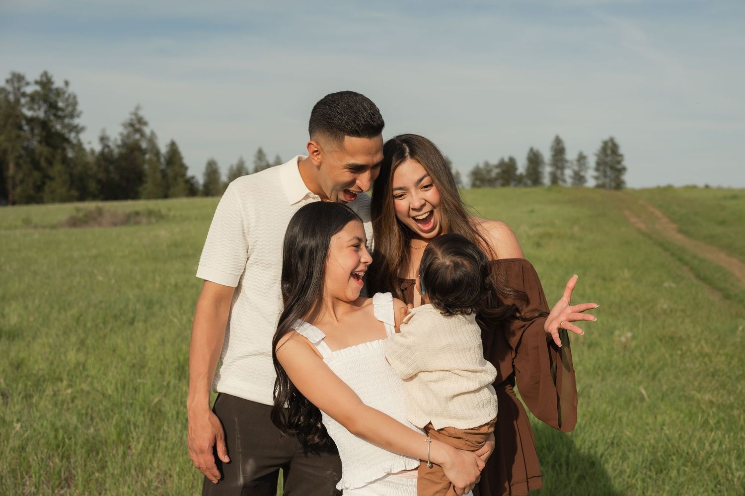 Happy family of four laughing together in a green meadow with trees in the background on a sunny day.