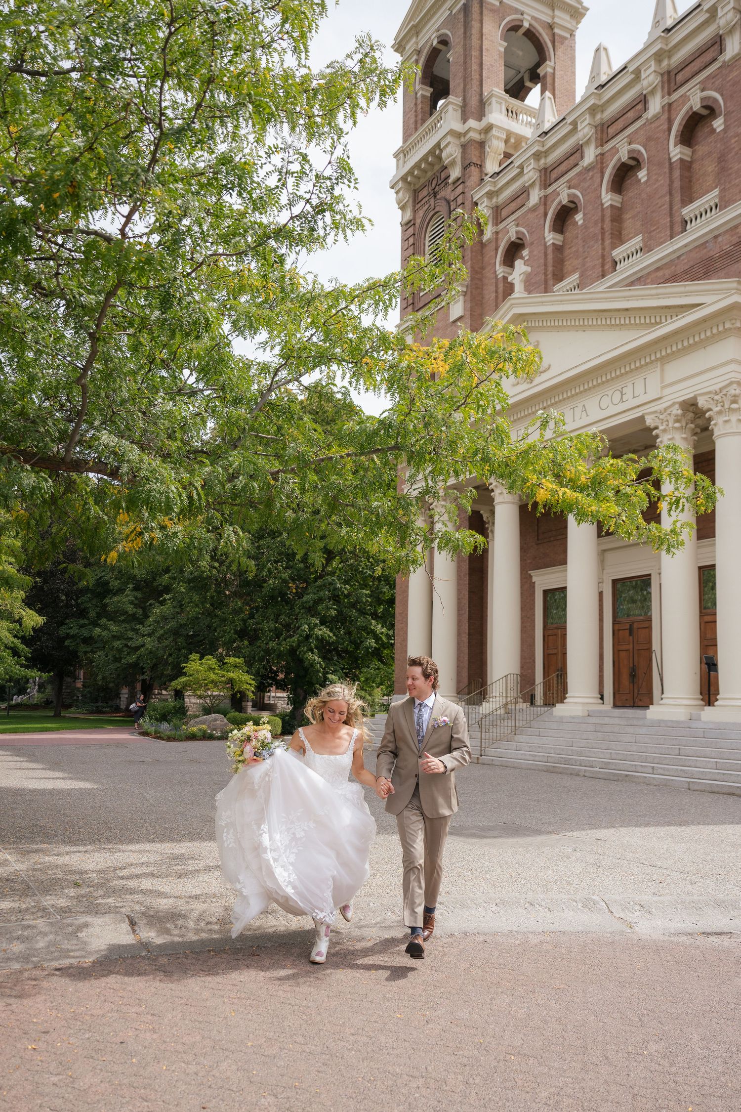 Bride and groom joyfully run hand-in-hand outside a grand brick church with tall bell tower on their wedding day.