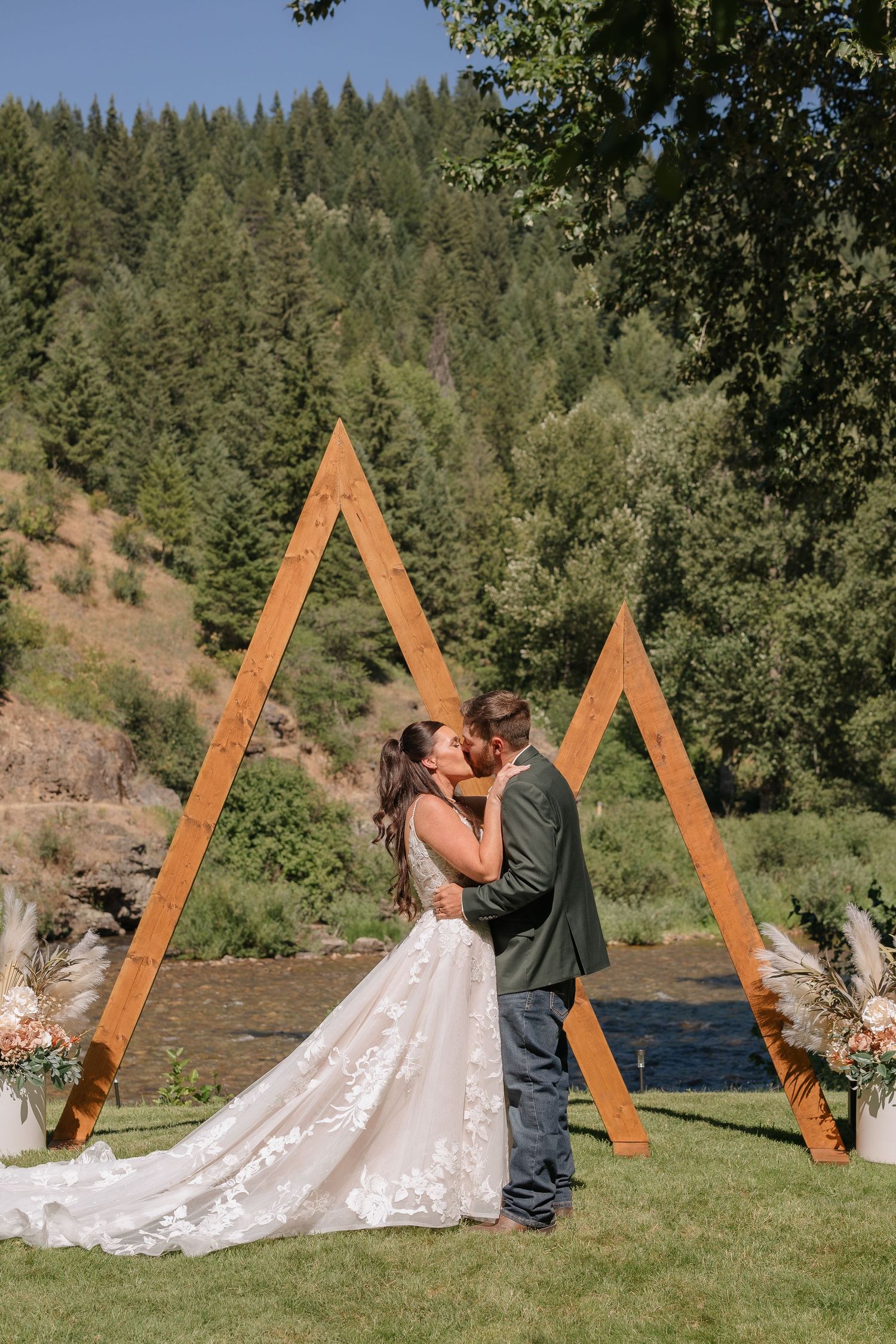 A couple shares a romantic moment in front of a triangular wooden arch during an outdoor wedding ceremony in a forest setting.