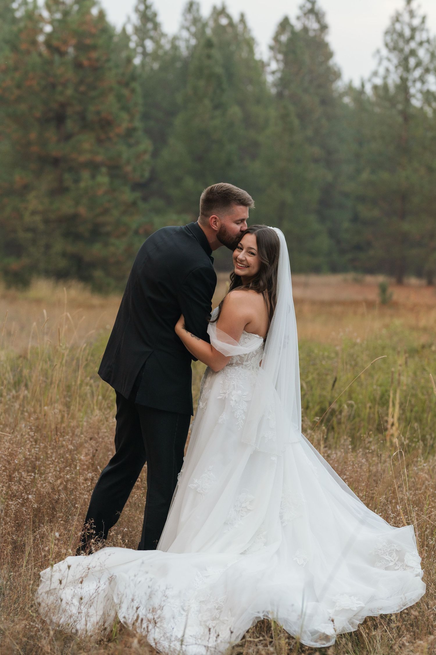 A romantic wedding couple embraces in a rustic field with evergreen trees in the background.