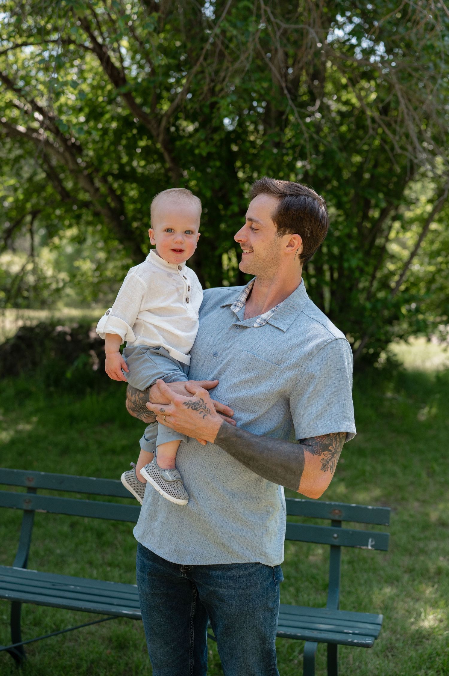 A playful moment captured outdoors on a park bench as an adult holds a small child against a leafy green background.