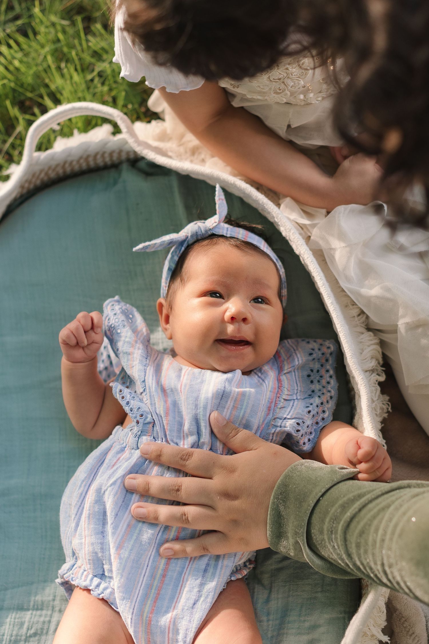 Happy baby in blue striped outfit with matching headband lying on teal cushion in garden setting.