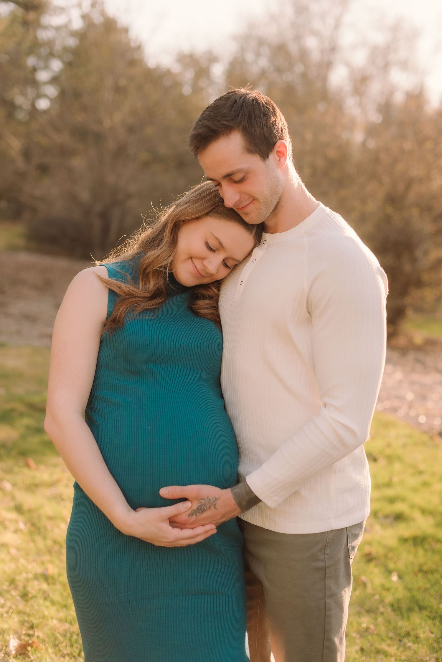 Couple shares intimate moment during maternity photoshoot at sunset in a grassy field.
