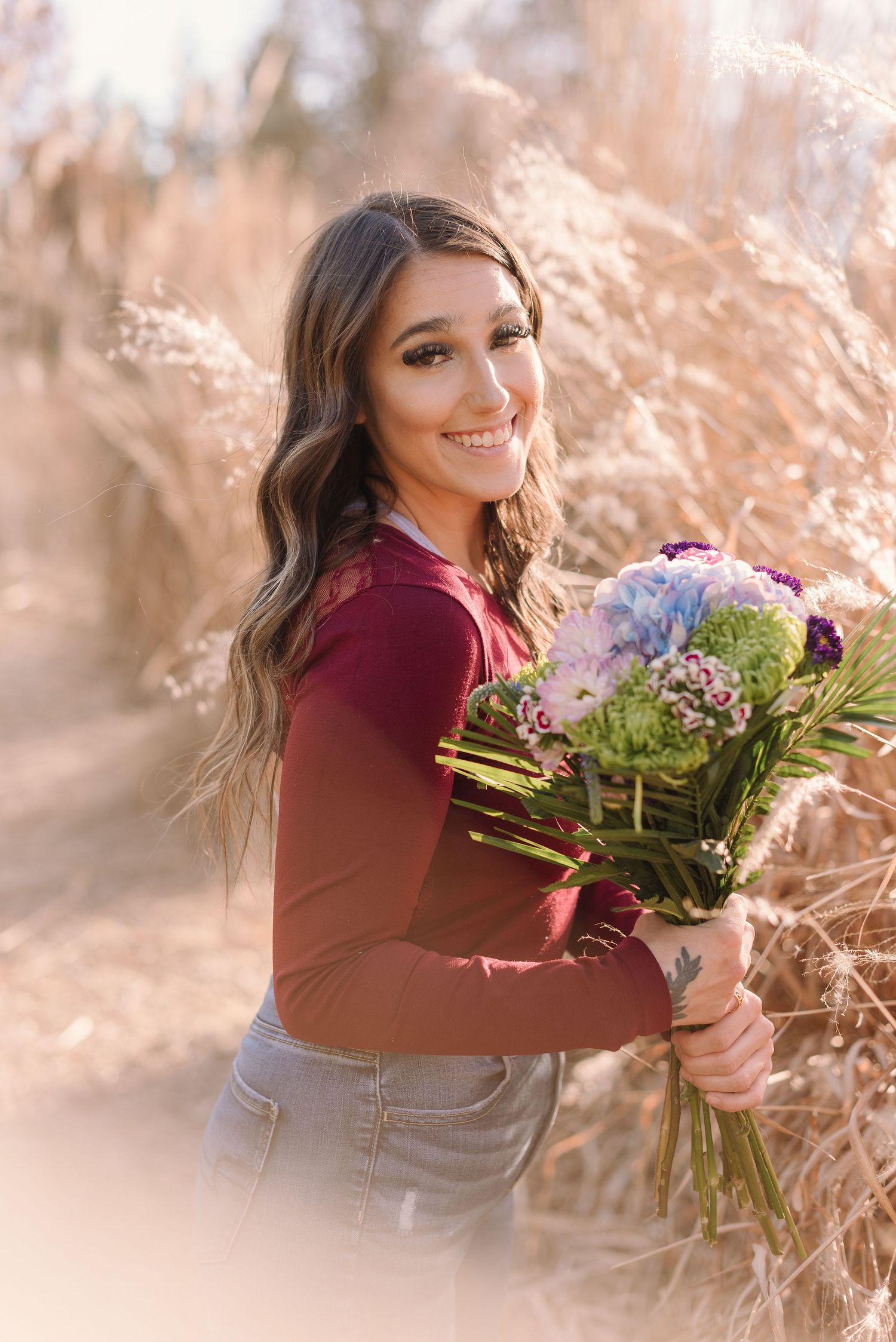A person in a burgundy sweater holds a vibrant bouquet of flowers while posing outdoors in warm autumn light.