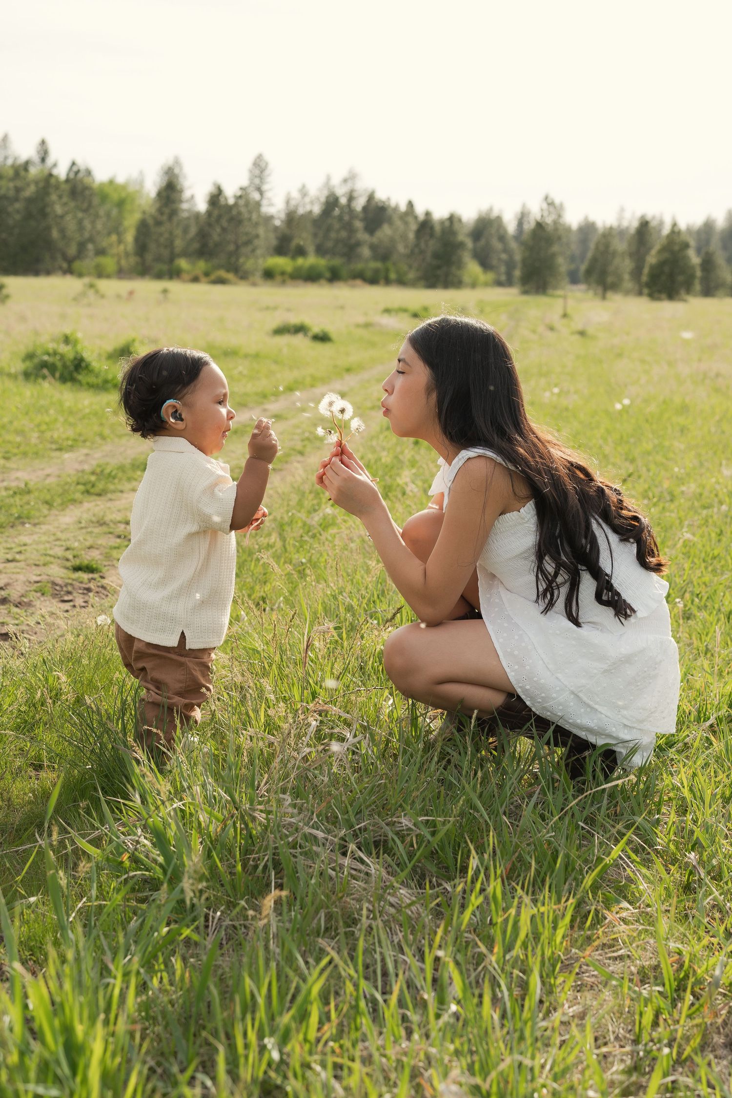 A mother and toddler blowing dandelion seeds together in a sunny green meadow with trees in the background.