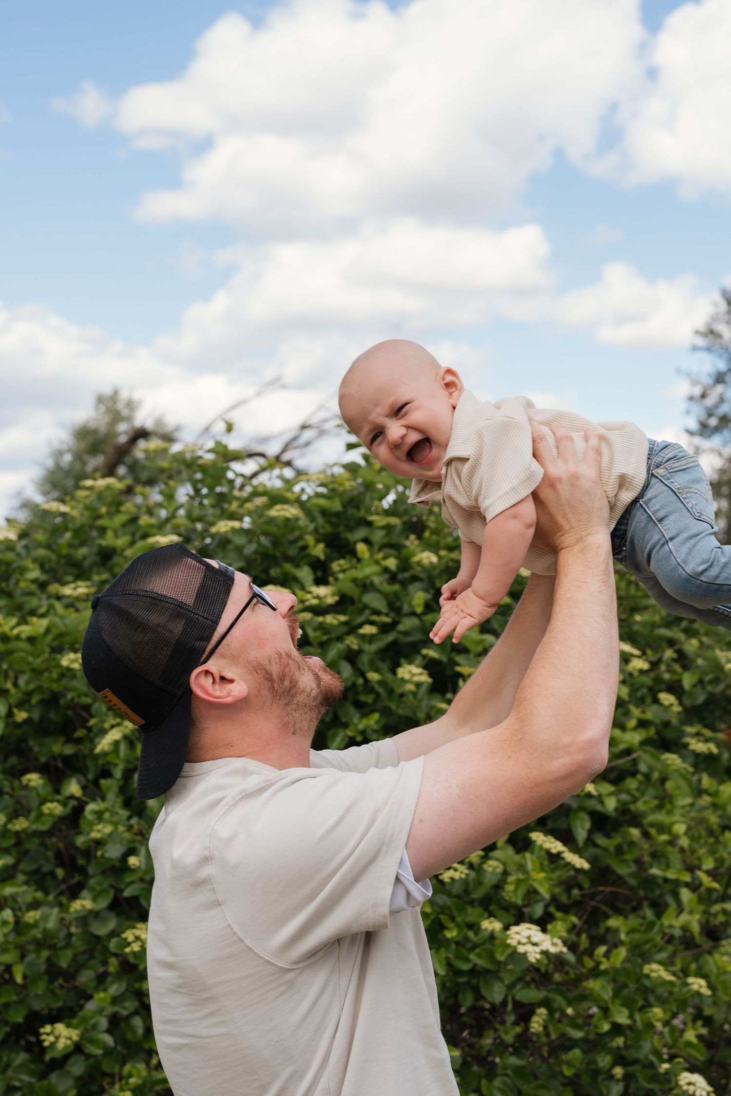 A joyful father lifts his laughing baby high in the air outdoors, surrounded by lush green trees under a cloudy sky.