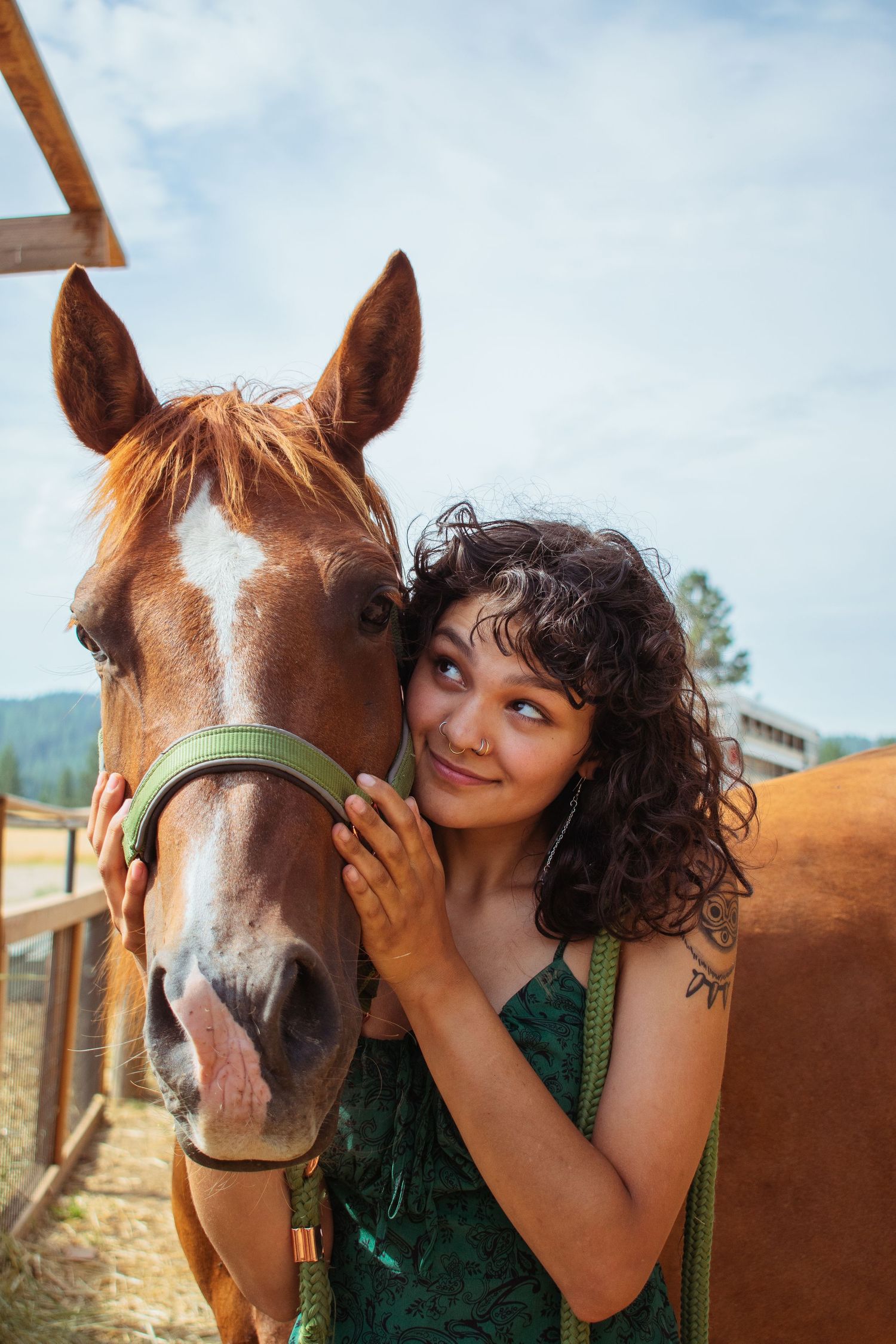 Young woman with curly hair tenderly embraces a chestnut horse at a sunny ranch, smiling warmly.