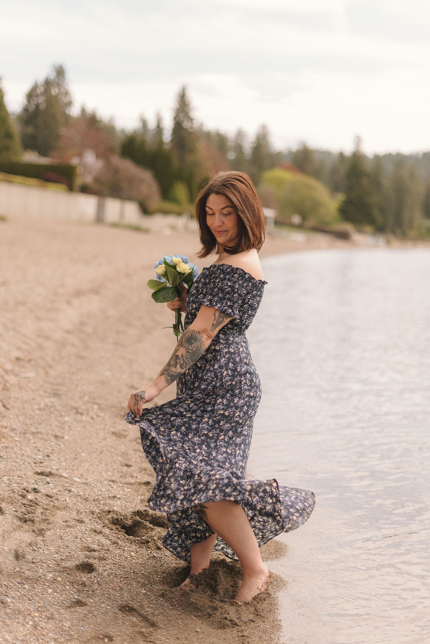 A person in a floral dress poses gracefully by the water's edge on a sandy beach with trees in the background.