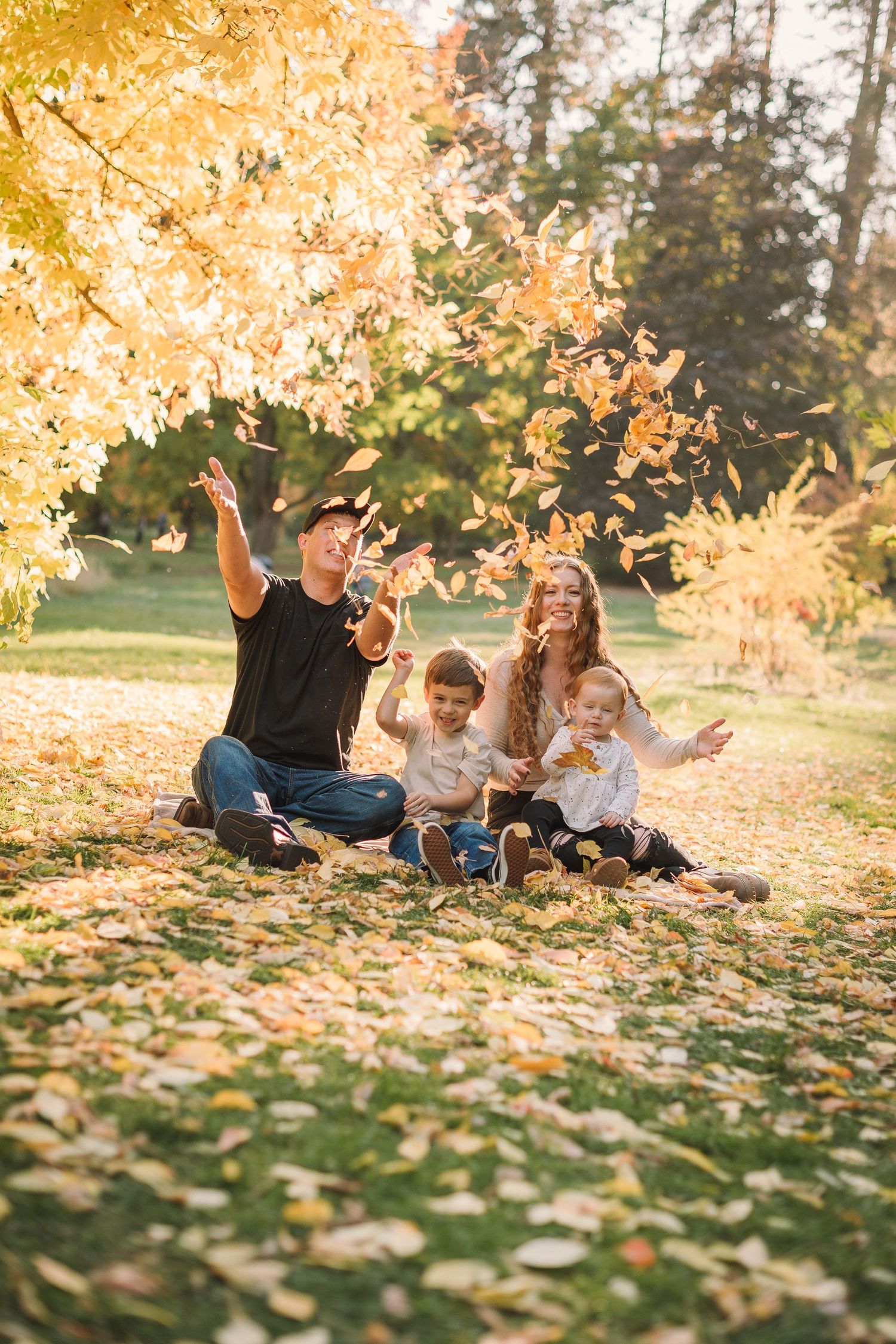 A family relaxes together on fallen autumn leaves during a golden sunset in a park setting.
