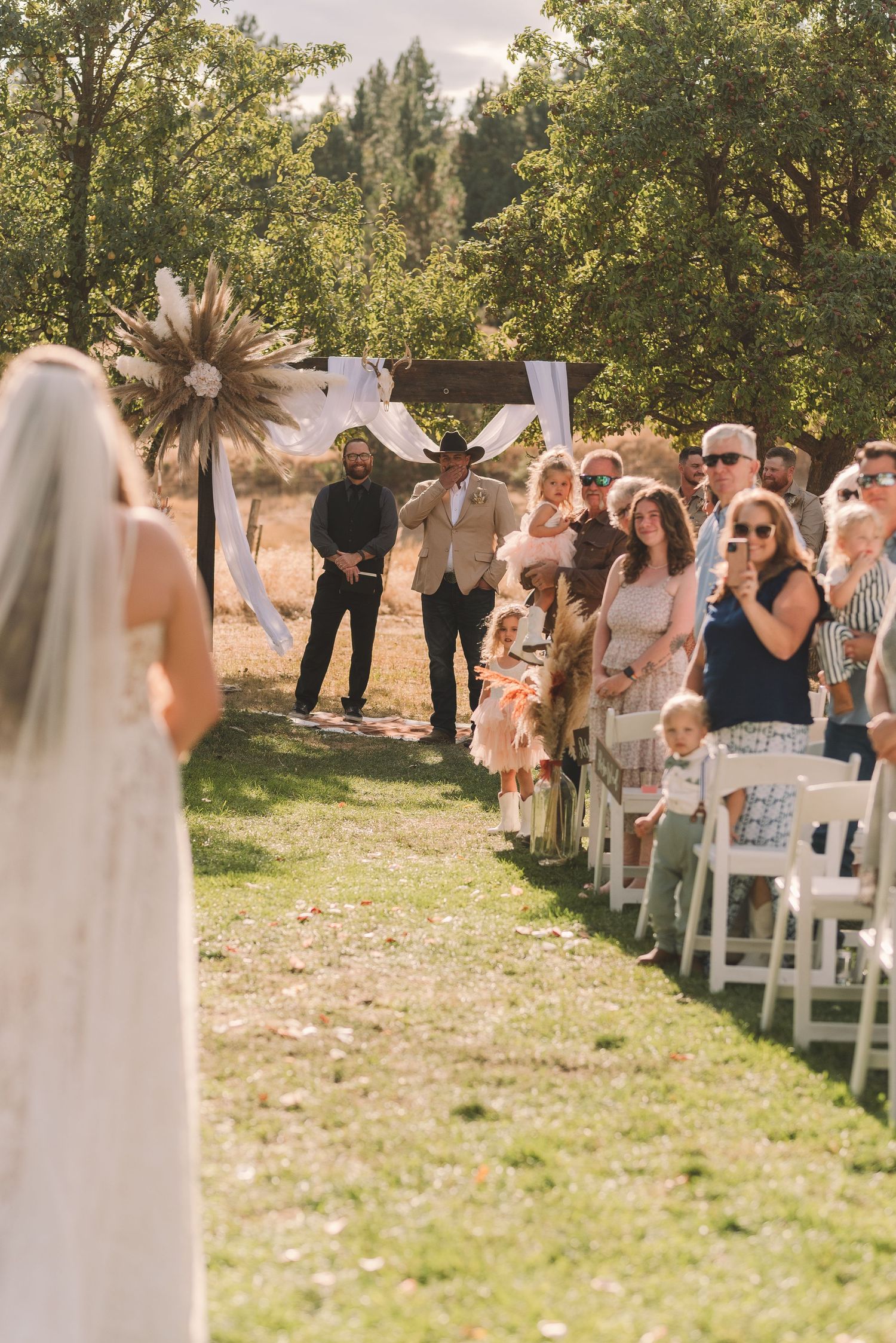 Wedding guests stand in rows of white chairs on a grassy lawn during an outdoor ceremony.