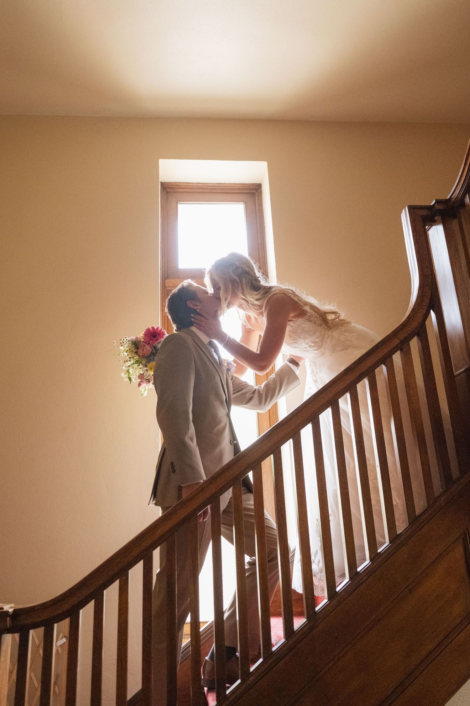 Bride and groom sharing a romantic kiss on a sunlit staircase, groom holding a colorful bouquet in a warmly lit interior.