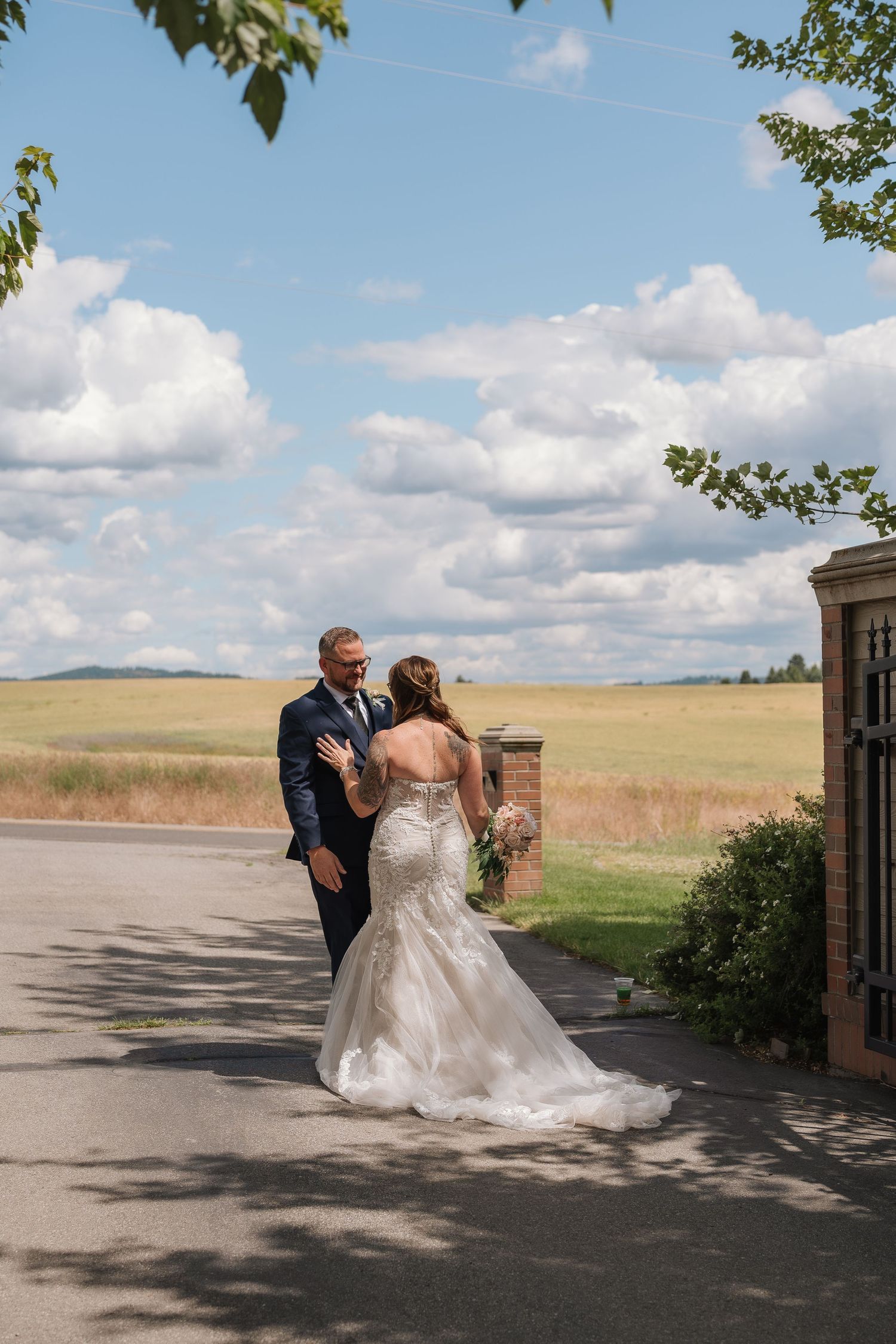 A couple in formal wedding attire dances together on a paved area with rolling hills and blue sky in the background.