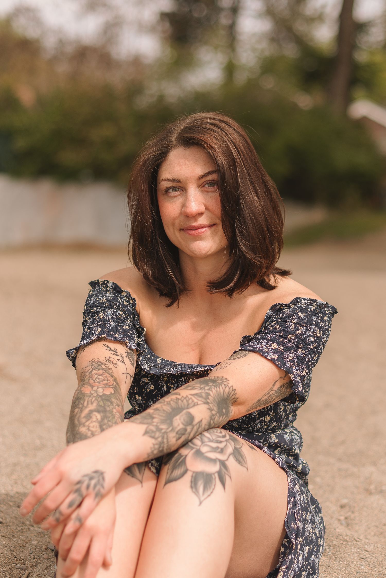 Person wearing a floral dress sits on sandy beach showing arm tattoos and relaxed pose in natural lighting.