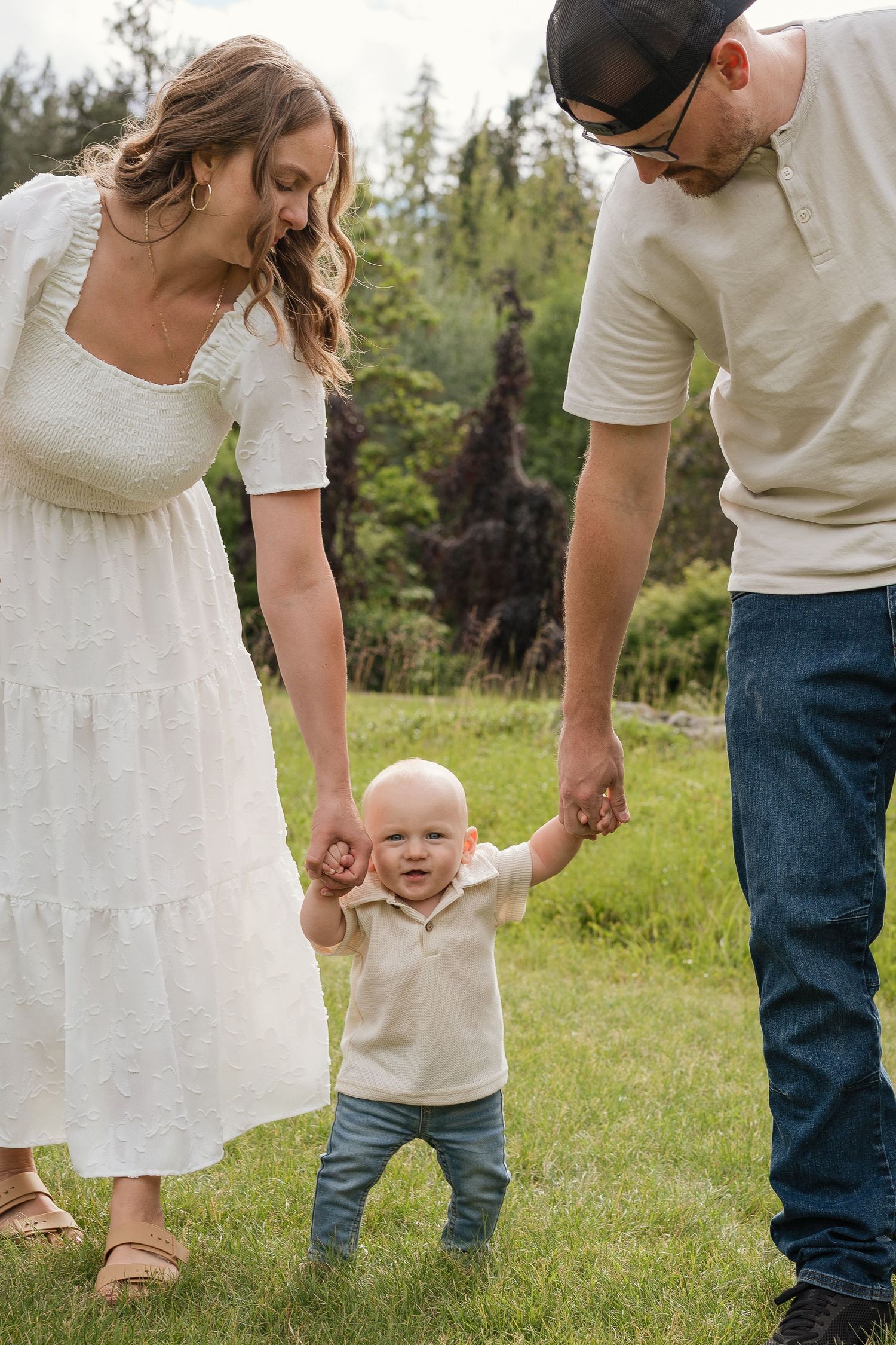 A young family helps their smiling toddler take first steps on green grass outdoors.