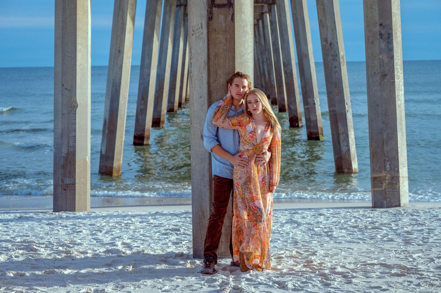 beautiful active, boho, engaged couple embracing against beach pier backdrop under sunset skies on sandy beach, editorial