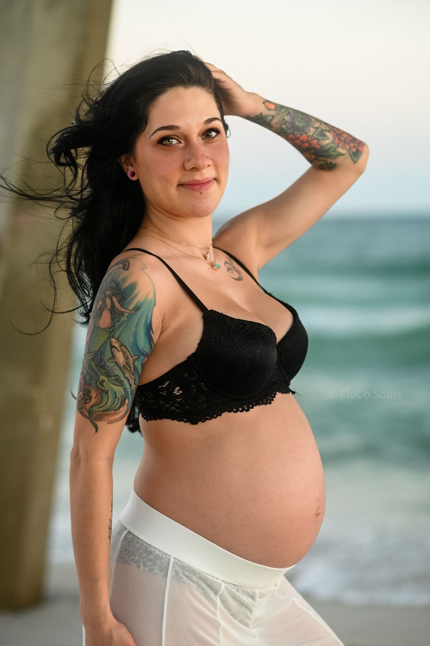 beautiful tattooed pregnant woman leaning against beach pier with blue eyes black bra and free people skirt