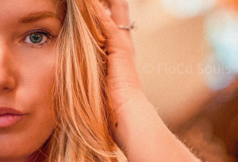 Cropped portrait showing green eye and blonde hair against a warm-toned blurred bokeh background.