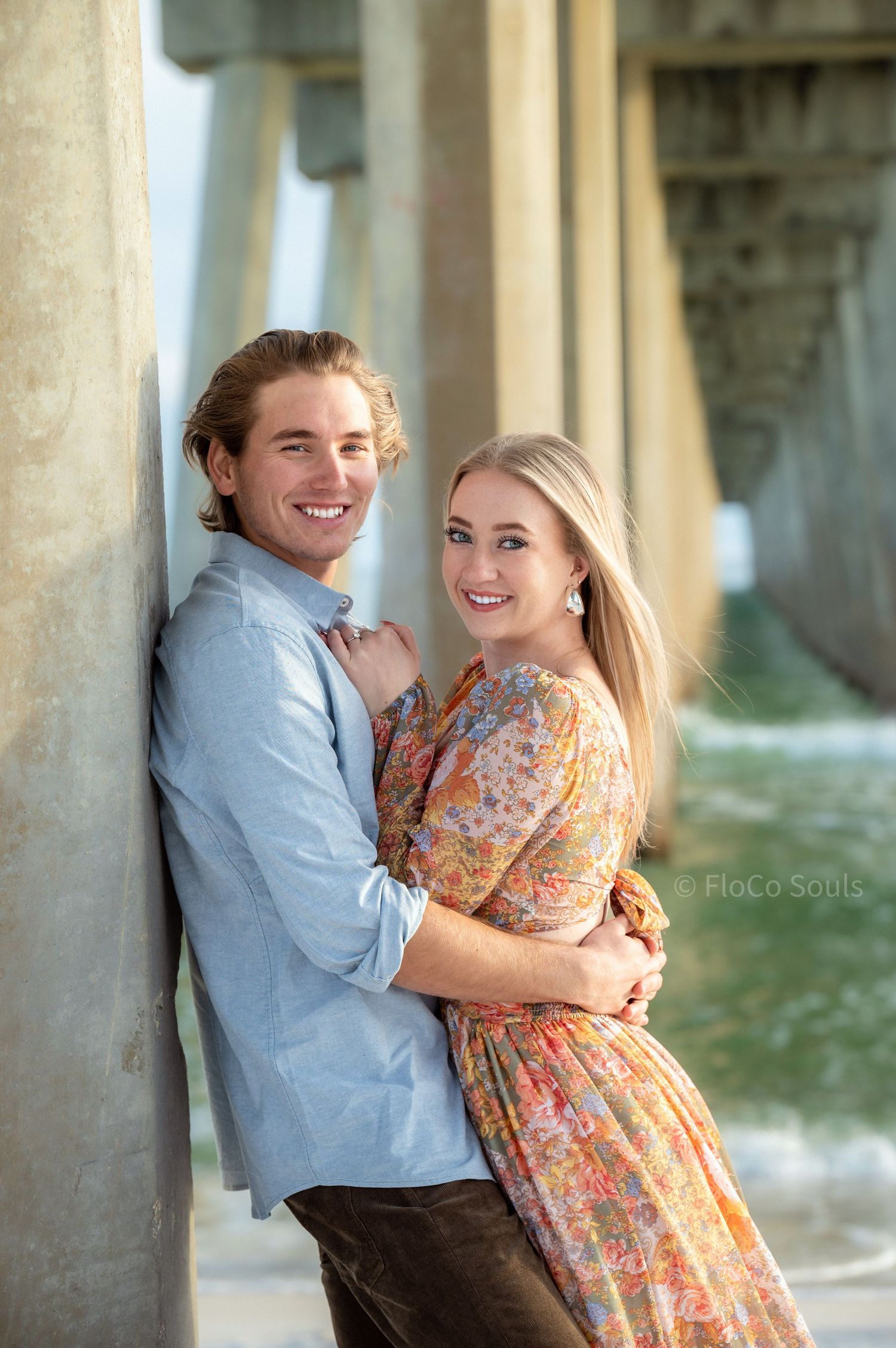 beautiful engaged couple under beach pier with dramatic waves, boho floral and blue linen