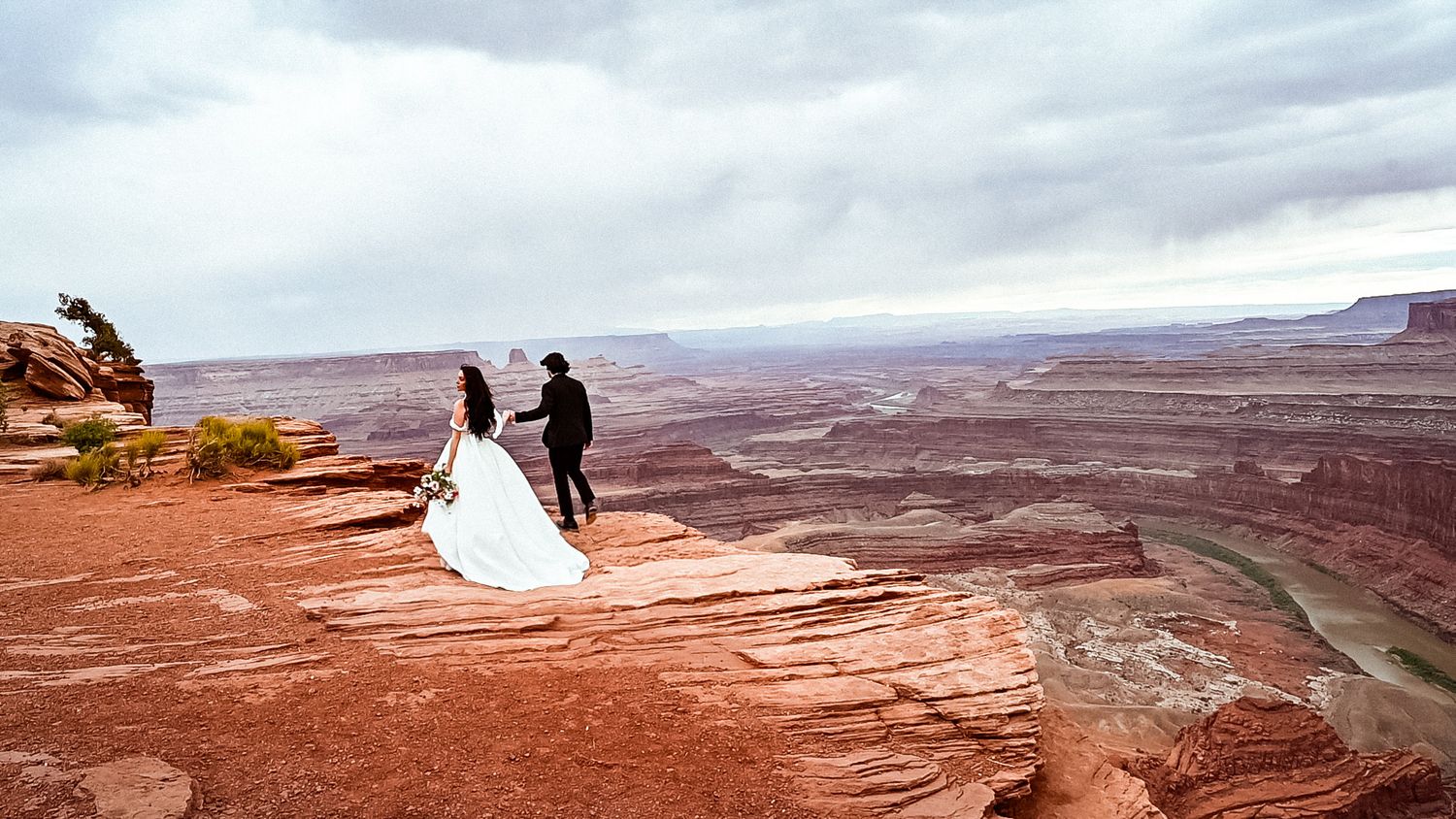 Bride & groom stand on a red sandstone cliff overlooking the vast desert canyons of Dead Horse Point State Park in Utah.