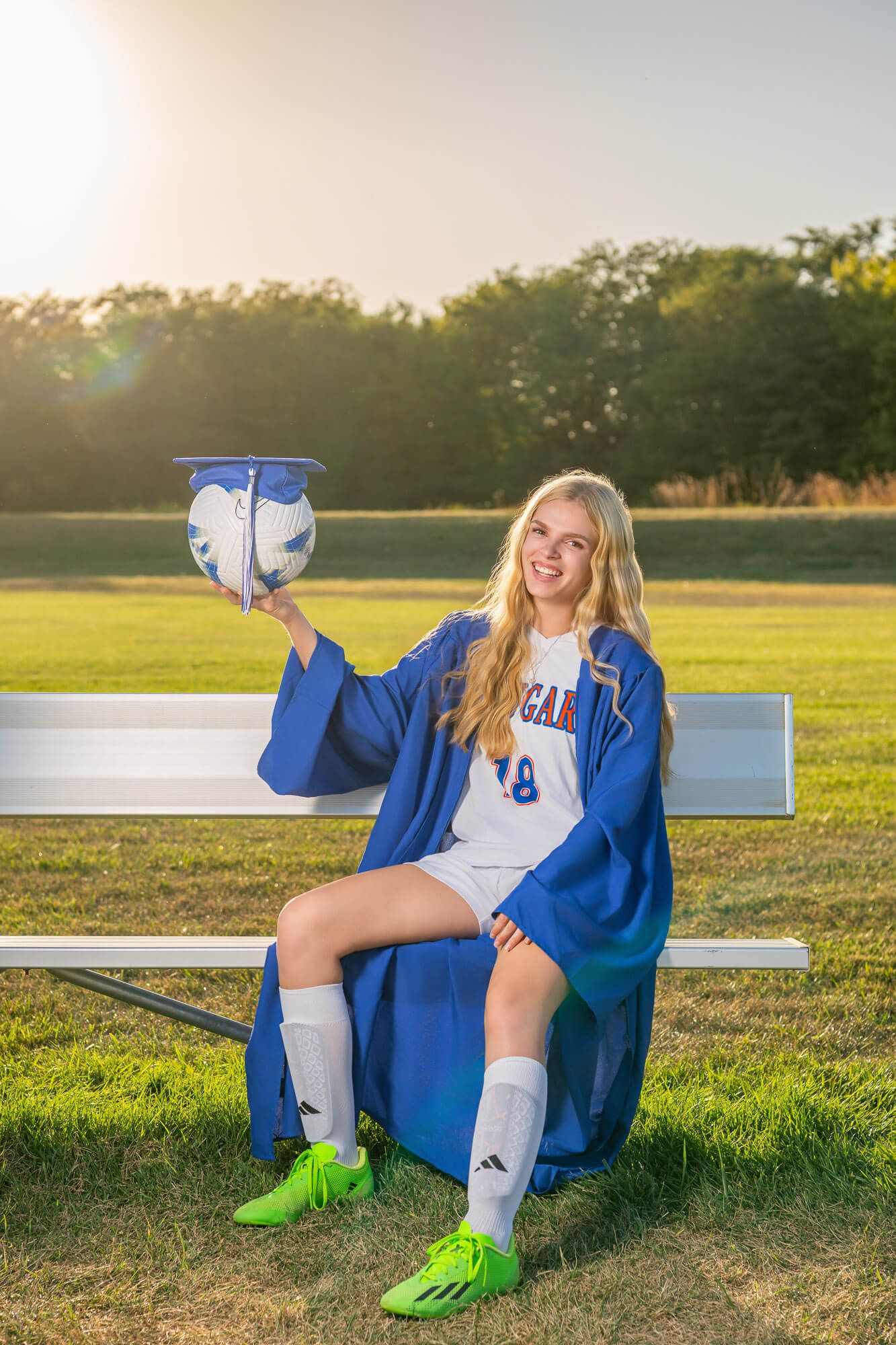 Blonde graduate in blue cap and gown holding soccer ball on field bench wearing green cleats and sports uniform.