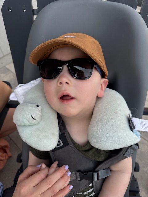 A child wearing sunglasses and a brown cap rests against a neck pillow while seated in a chair.