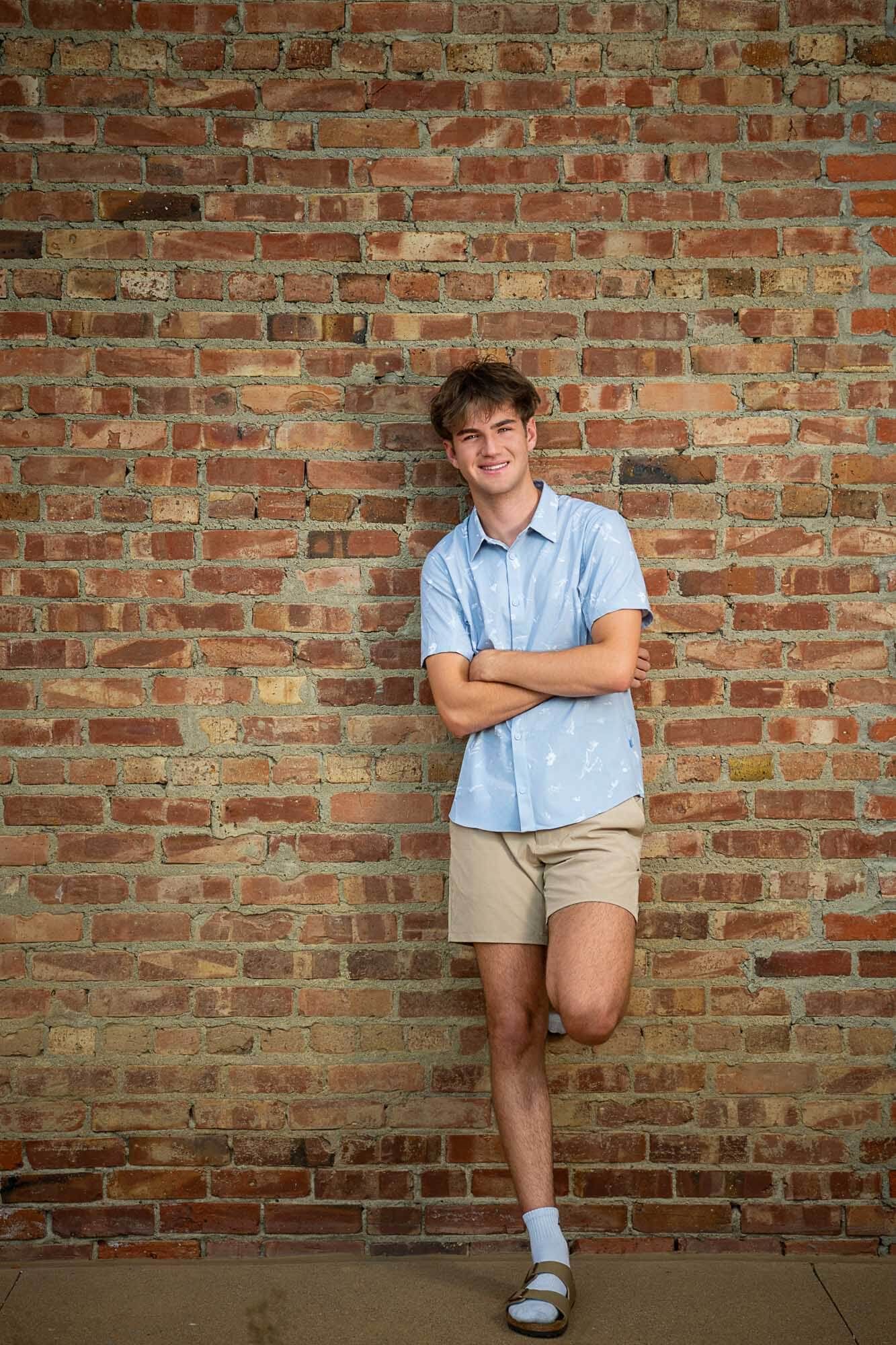 A casual portrait against a rustic brick wall wearing a light blue shirt and khaki shorts.
