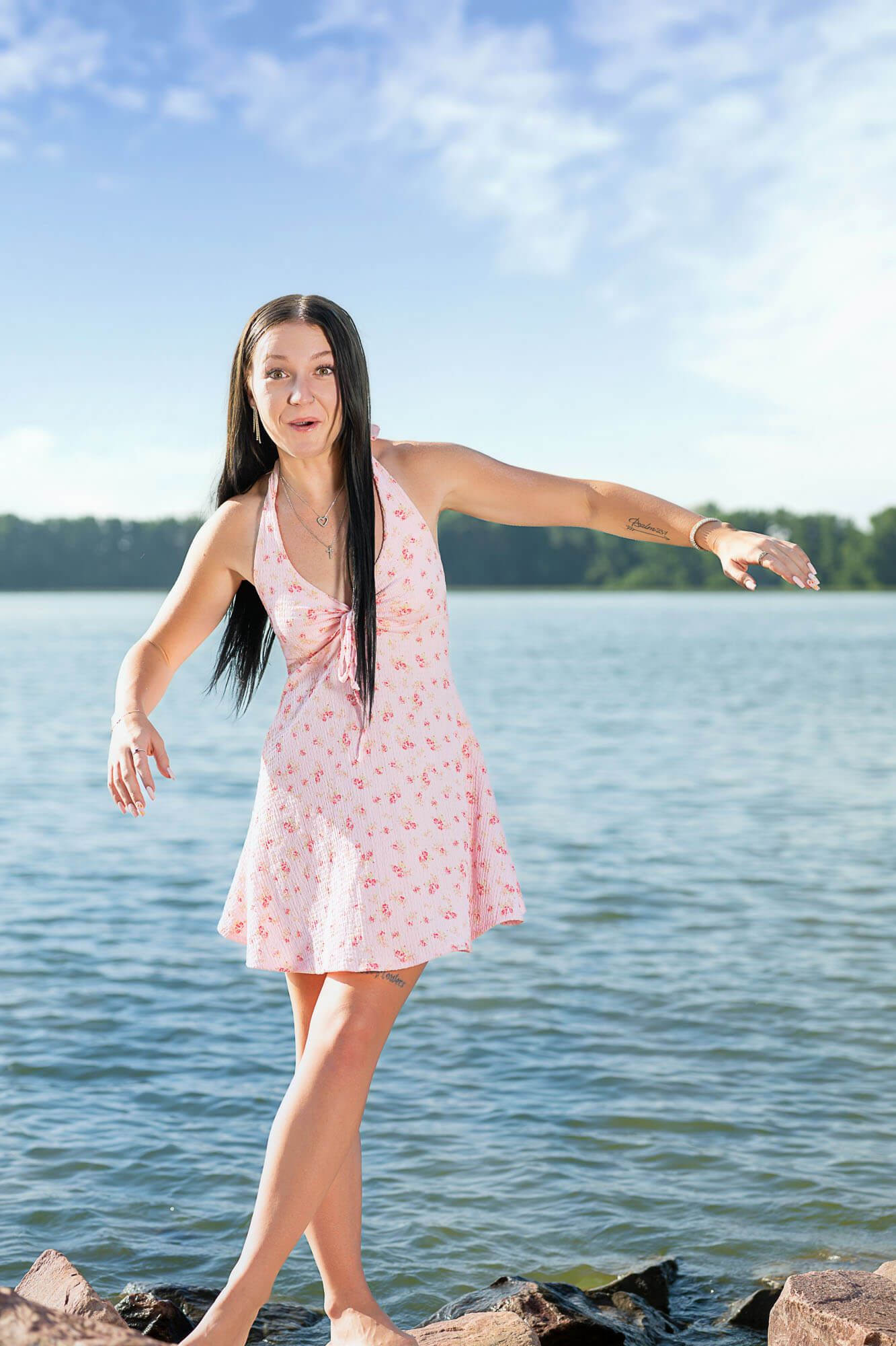 A person in a flowing pink dress strikes dynamic poses by a lake on a sunny day.