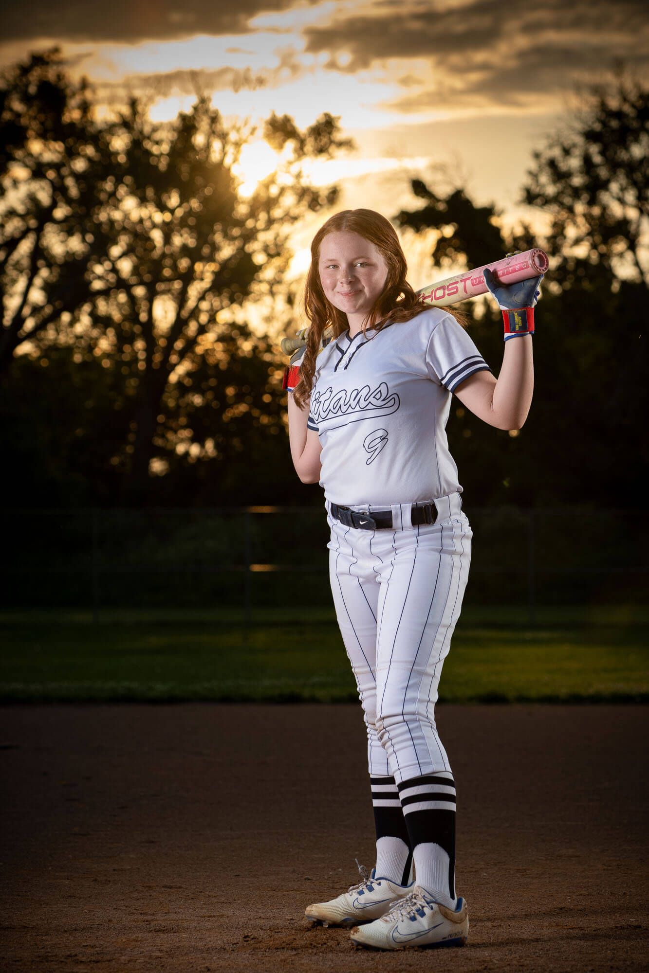 Young softball player in white uniform holding pink bat at sunset on dirt field.