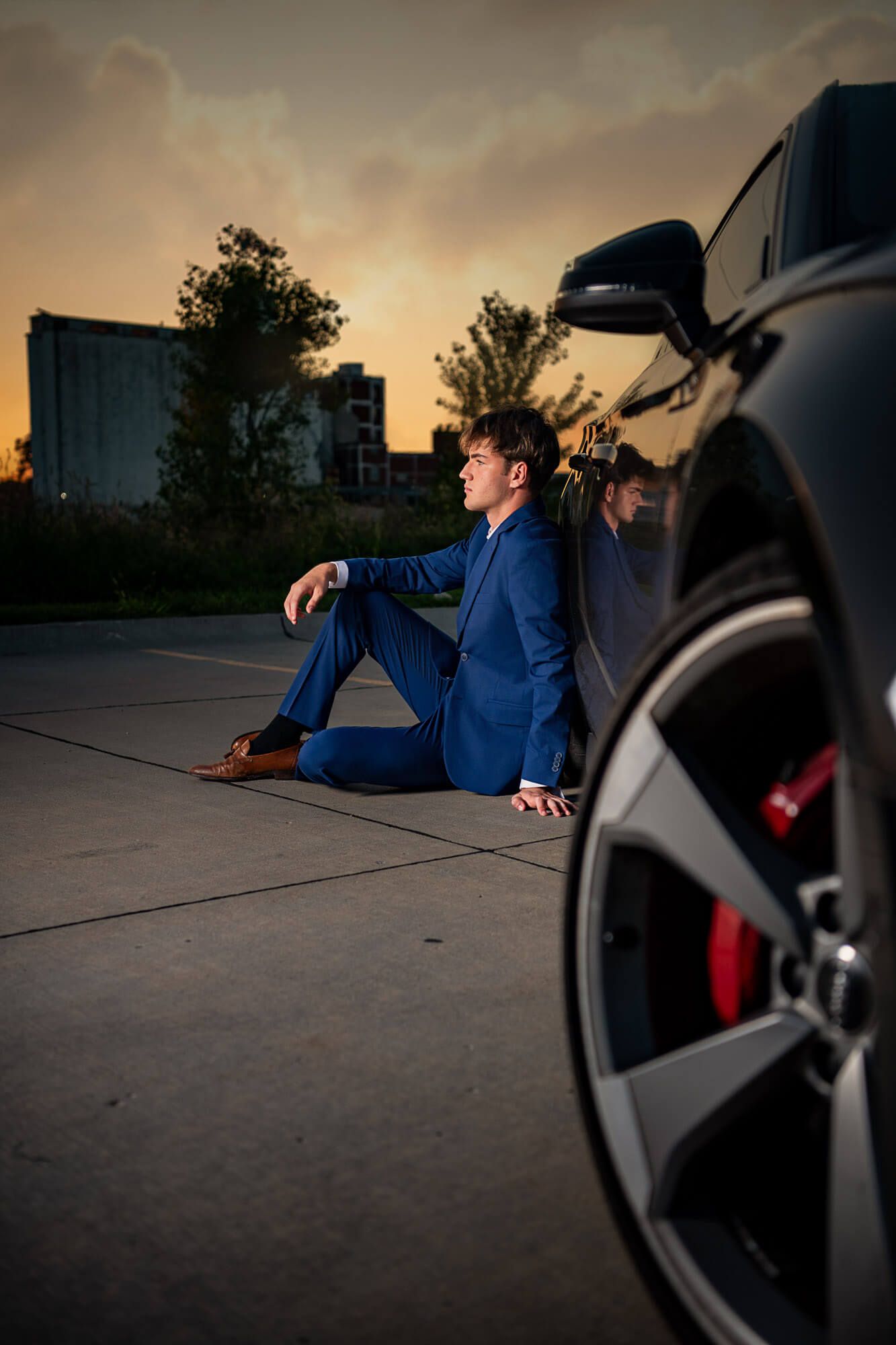 Senior photography session in Downtown Omaha of a male in a blue suit at sunset sitting down and leaning against his Audi with caliper shown