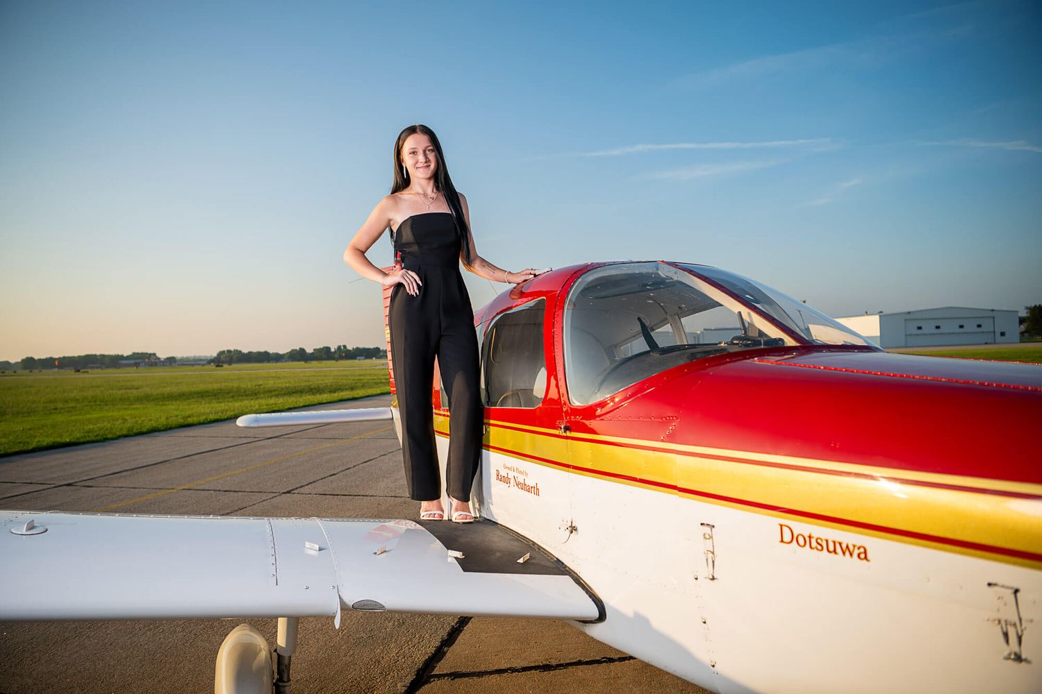 A small yellow and red private aircraft sits on a sunny runway with a figure standing beside it.