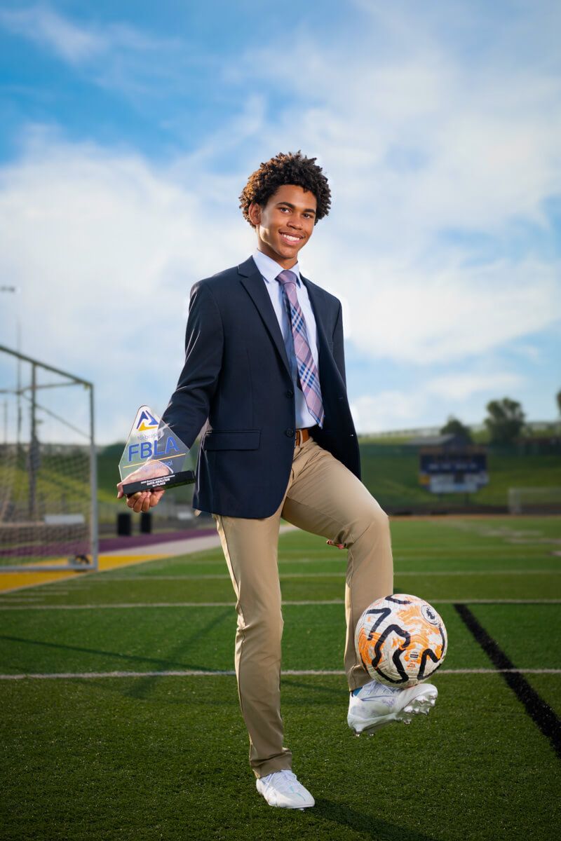 A smiling young man in a suit poses on a soccer field with a ball at his foot and an award in hand.