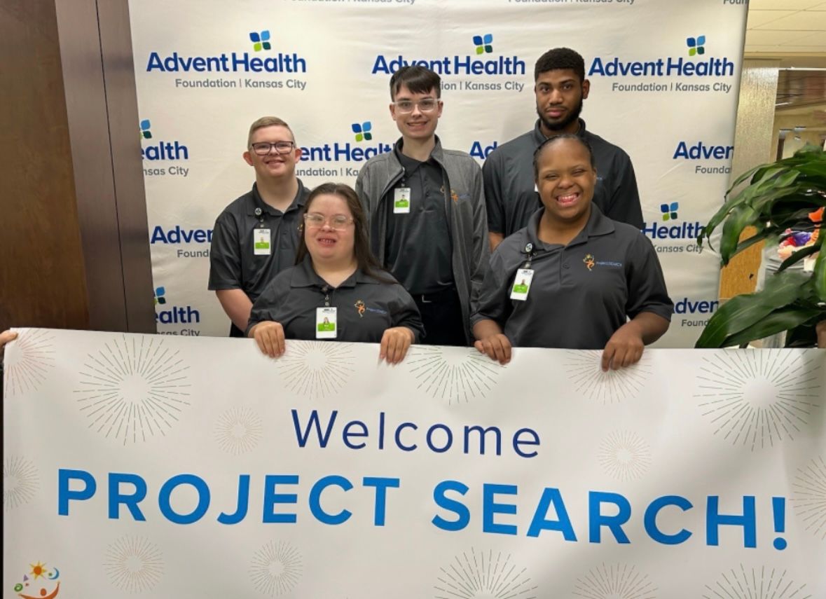 A group of employees pose together in front of an AdventHealth Project Search welcome banner and logo wall.