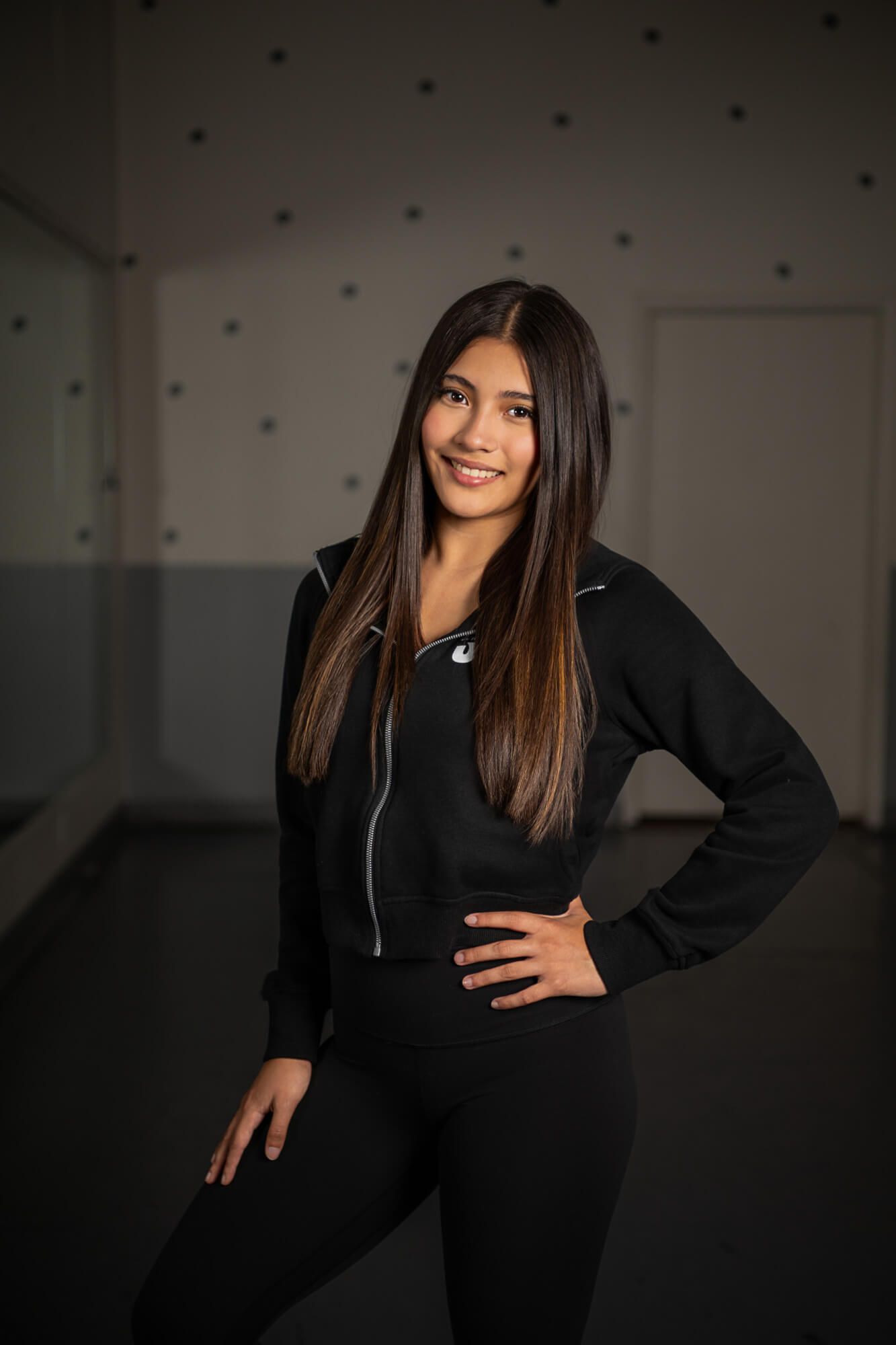 Athletic woman in all-black workout attire poses confidently against a dark wall with spotlights.